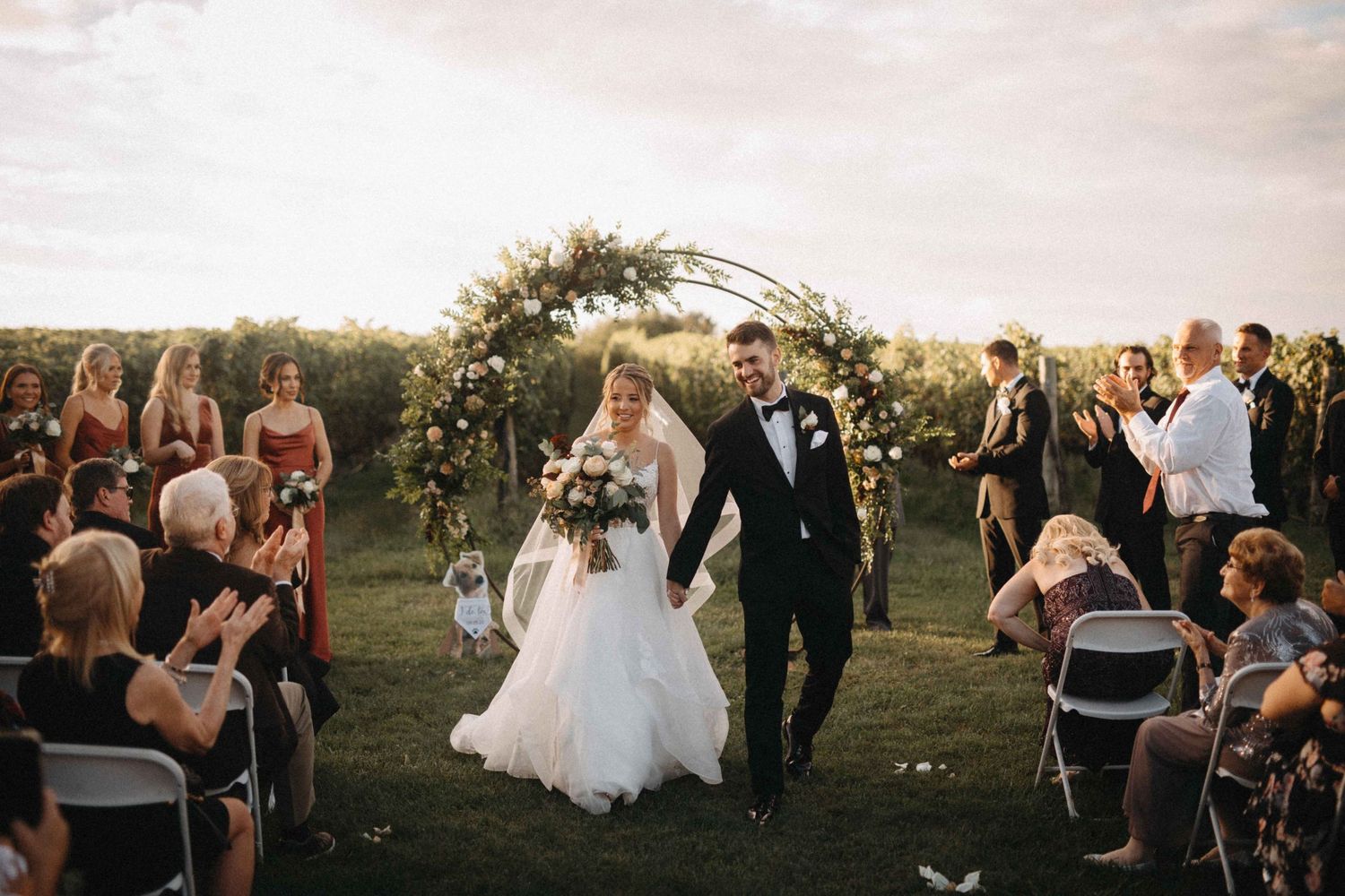 Newlyweds walk joyfully down aisle as guests applaud at outdoor vineyard wedding ceremony at sunset.