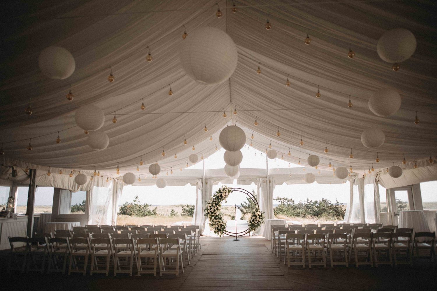 Stunning wedding tent ceremony space with white lanterns, draped ceiling, and floral arch.