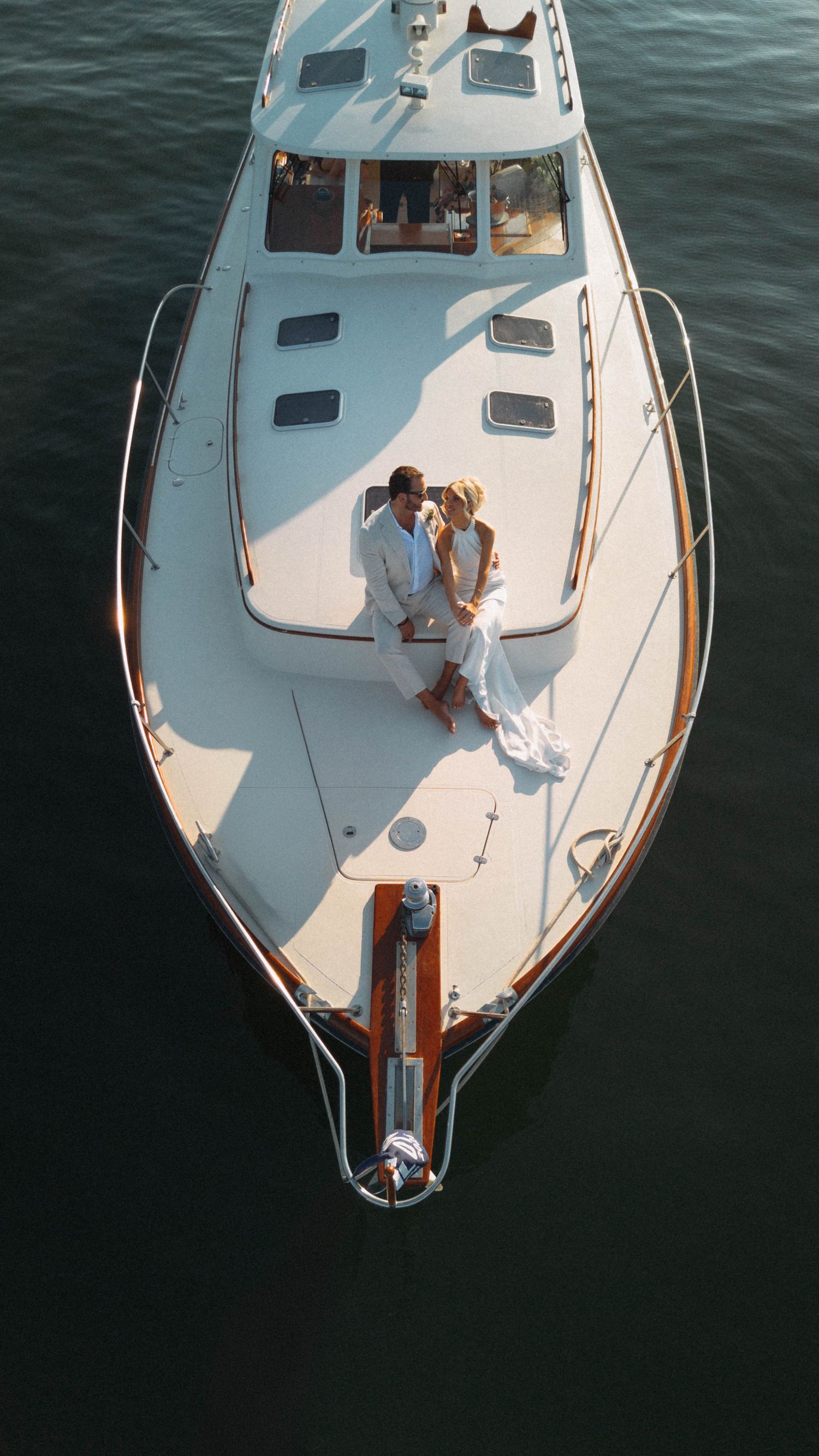 Aerial view of a bride and groom sitting together on the bow of a luxury white yacht.