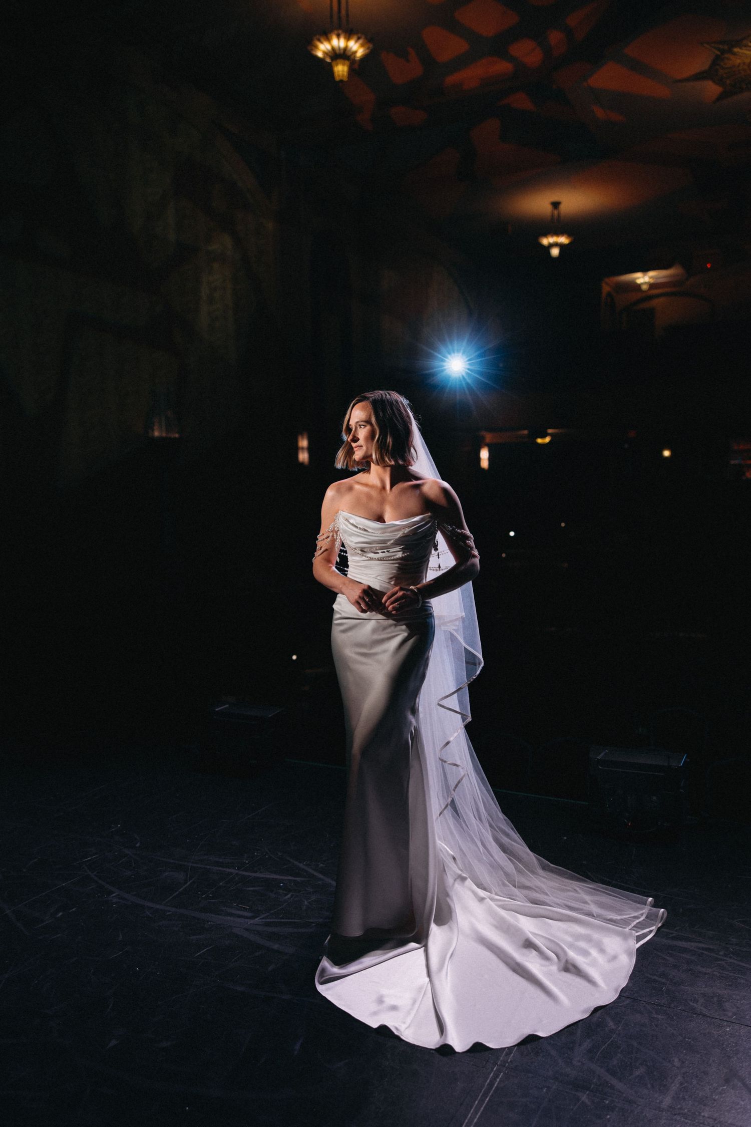 Elegant bride in off-shoulder white gown and veil poses dramatically in a dimly lit ornate venue.