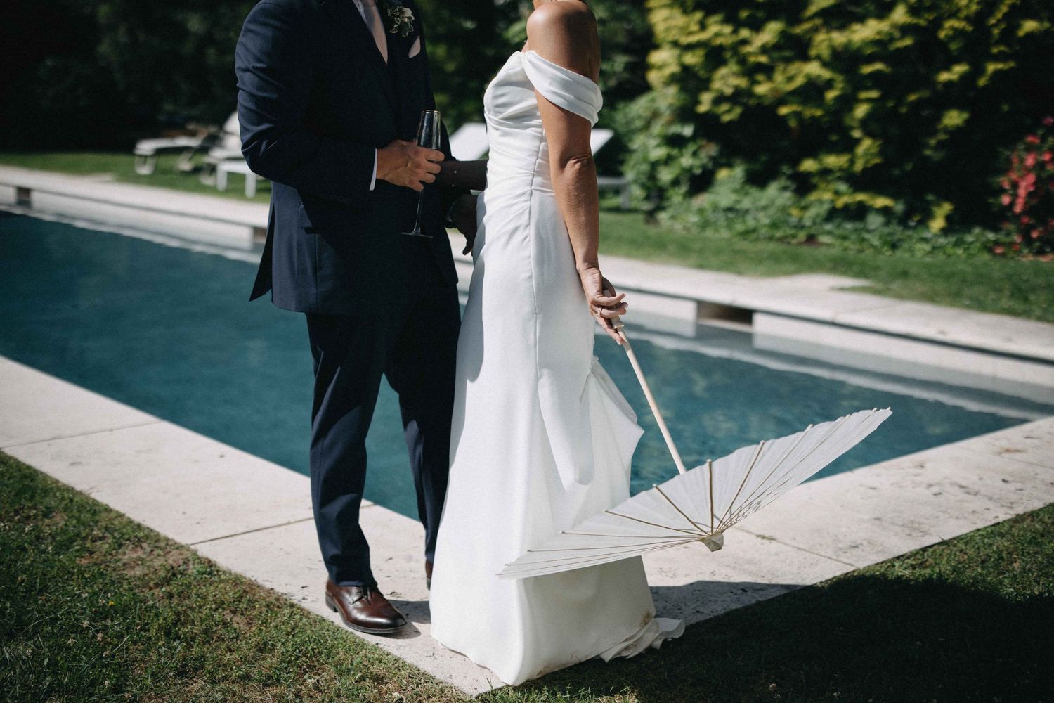 Bride in off-shoulder gown holds a white parasol beside her groom near a serene poolside garden setting.