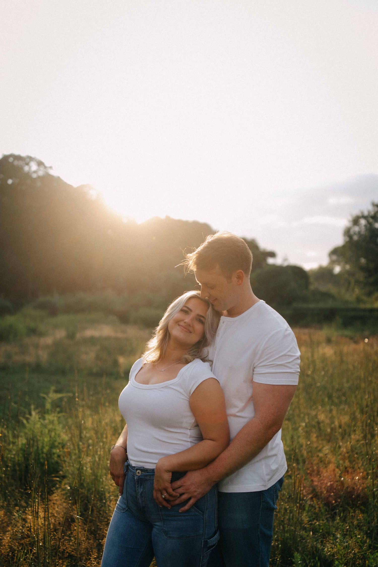 Couple embracing in a sunlit field at golden hour, woman smiling while leaning into her partner.