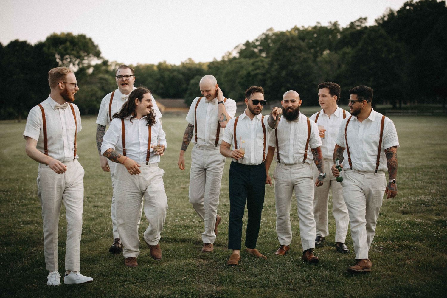 Groomsmen in white shirts and suspenders walk together outdoors on a green lawn during a wedding celebration.