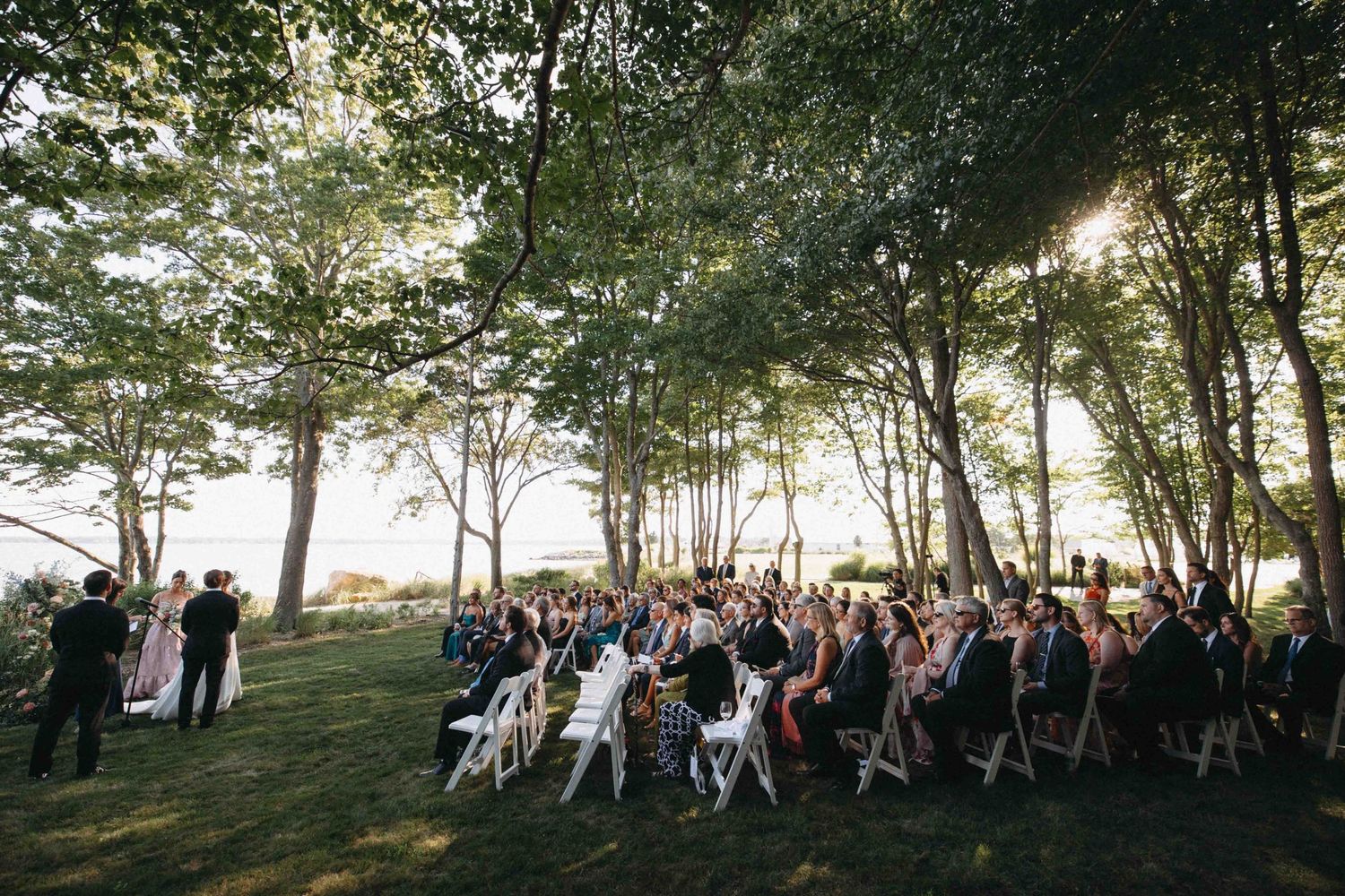 Outdoor wedding ceremony with guests seated on white chairs beneath tall trees by waterfront.
