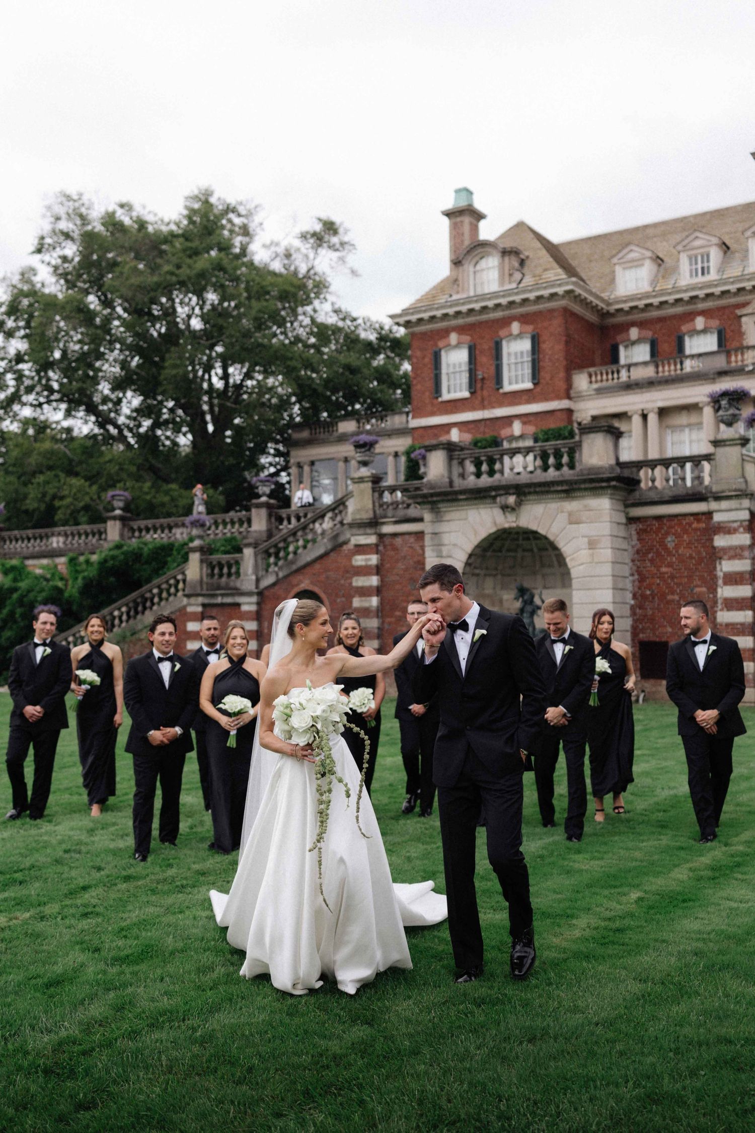 Bride and groom dance on green lawn before elegant brick mansion, wedding party in black attire stands behind them.