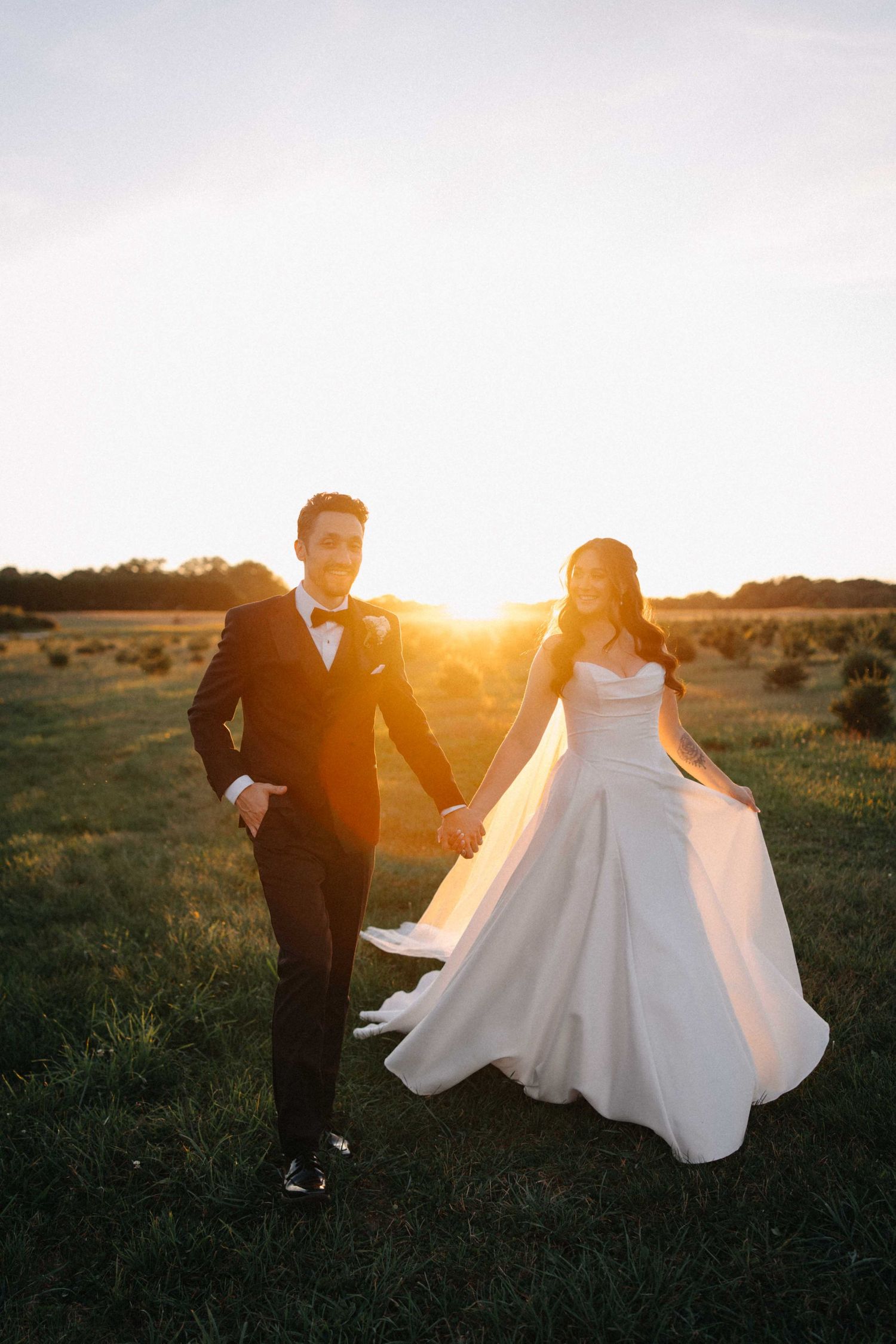 Bride and groom holding hands walking through golden field at stunning sunset.