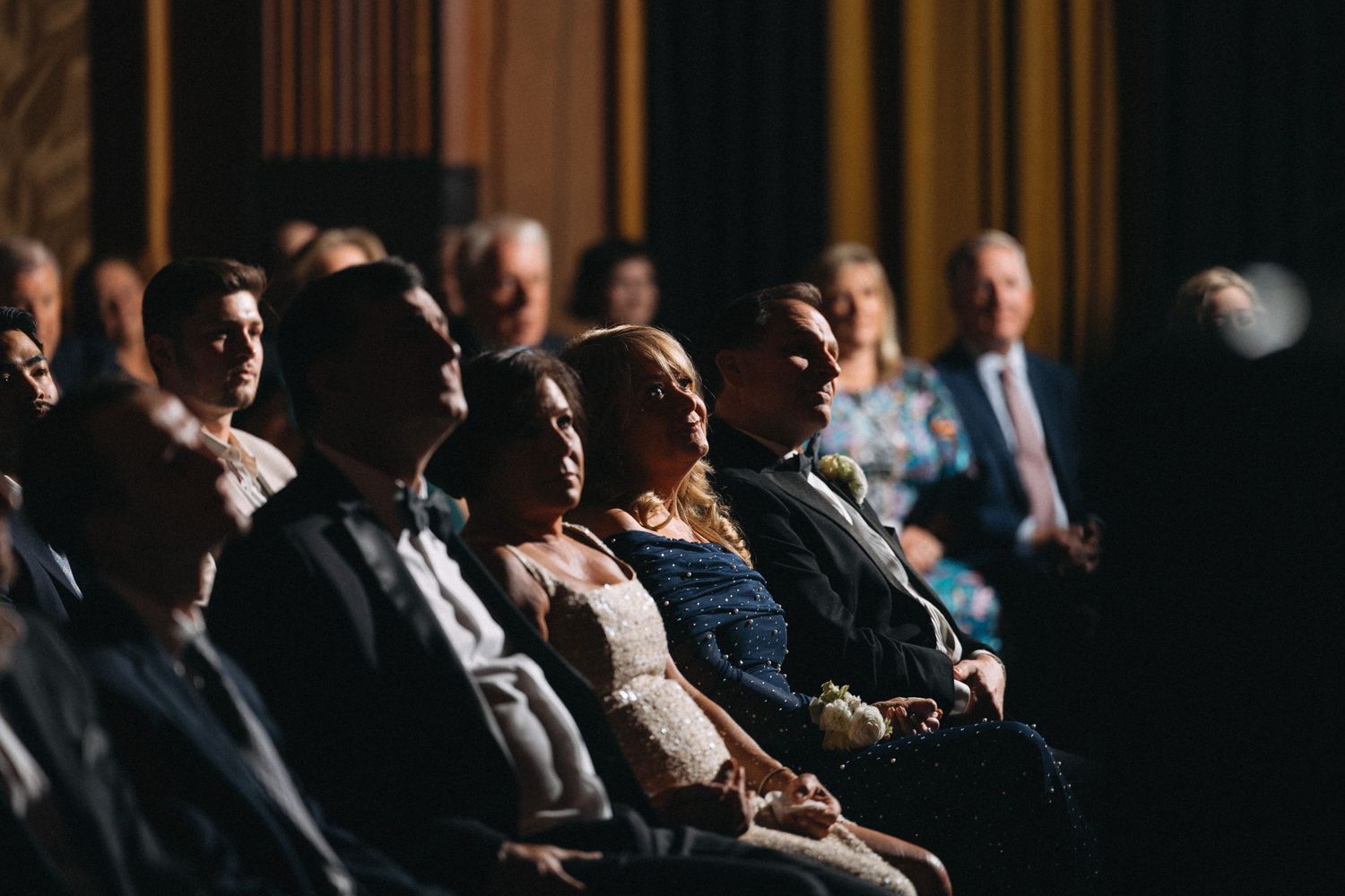 Wedding guests seated in rows enjoy the ceremony in a softly lit elegant venue with warm ambient lighting.