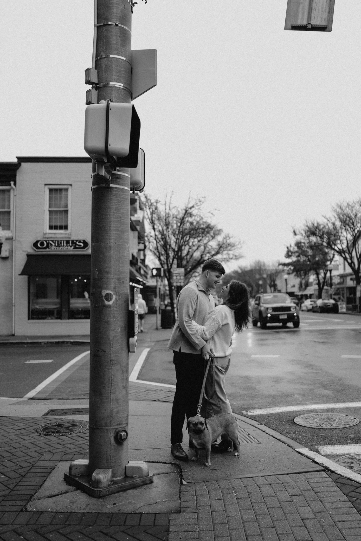 Black and white photo of a couple embracing at a city street corner with their dog nearby.