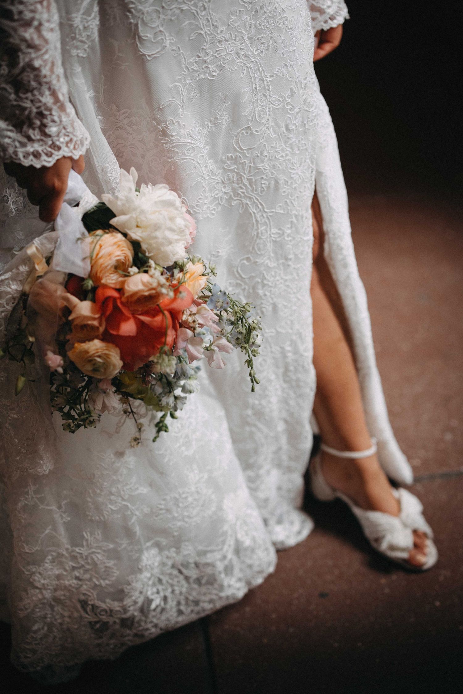 Close-up of a bride in a lace wedding dress holding a colorful bouquet with white sandals.