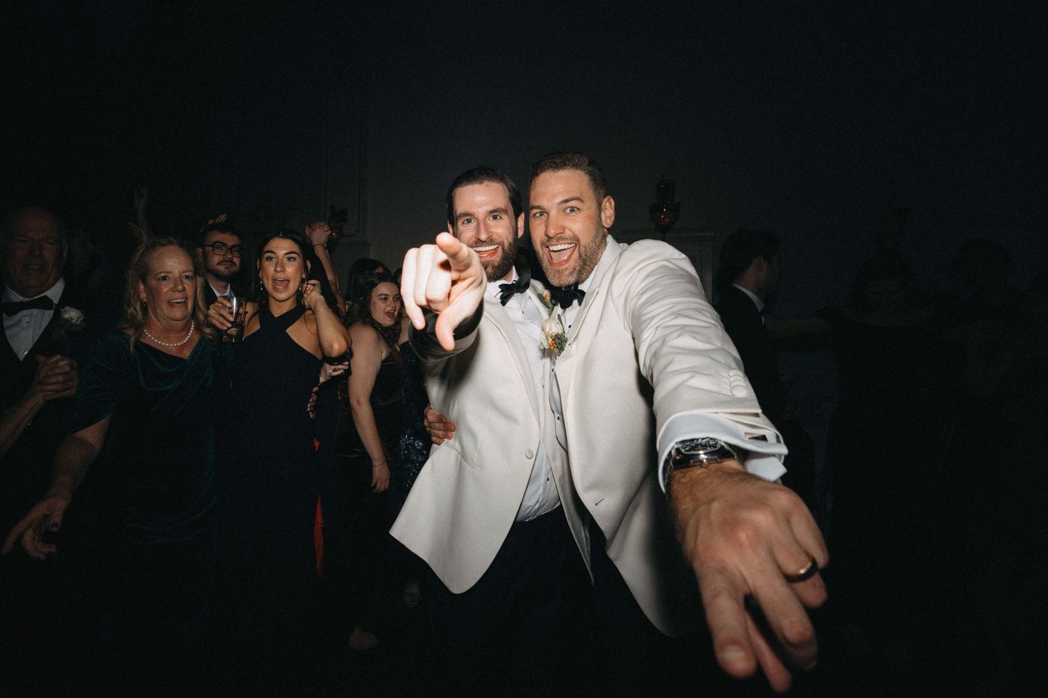 Groom in white tuxedo jacket celebrating with guests on wedding reception dance floor.