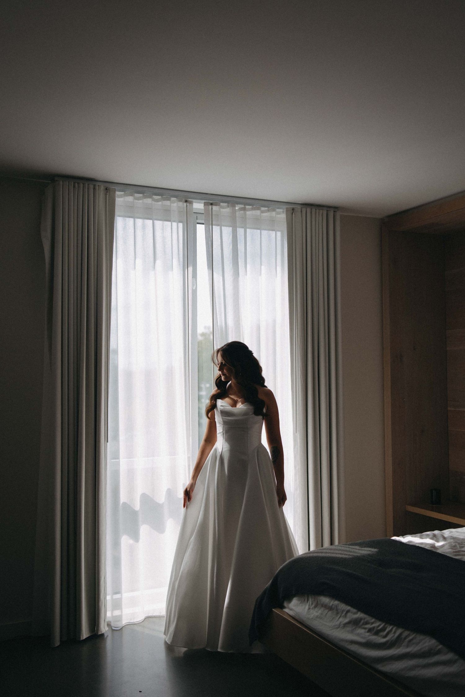 Bride in elegant white ball gown standing by sheer curtained window in dimly lit hotel room.