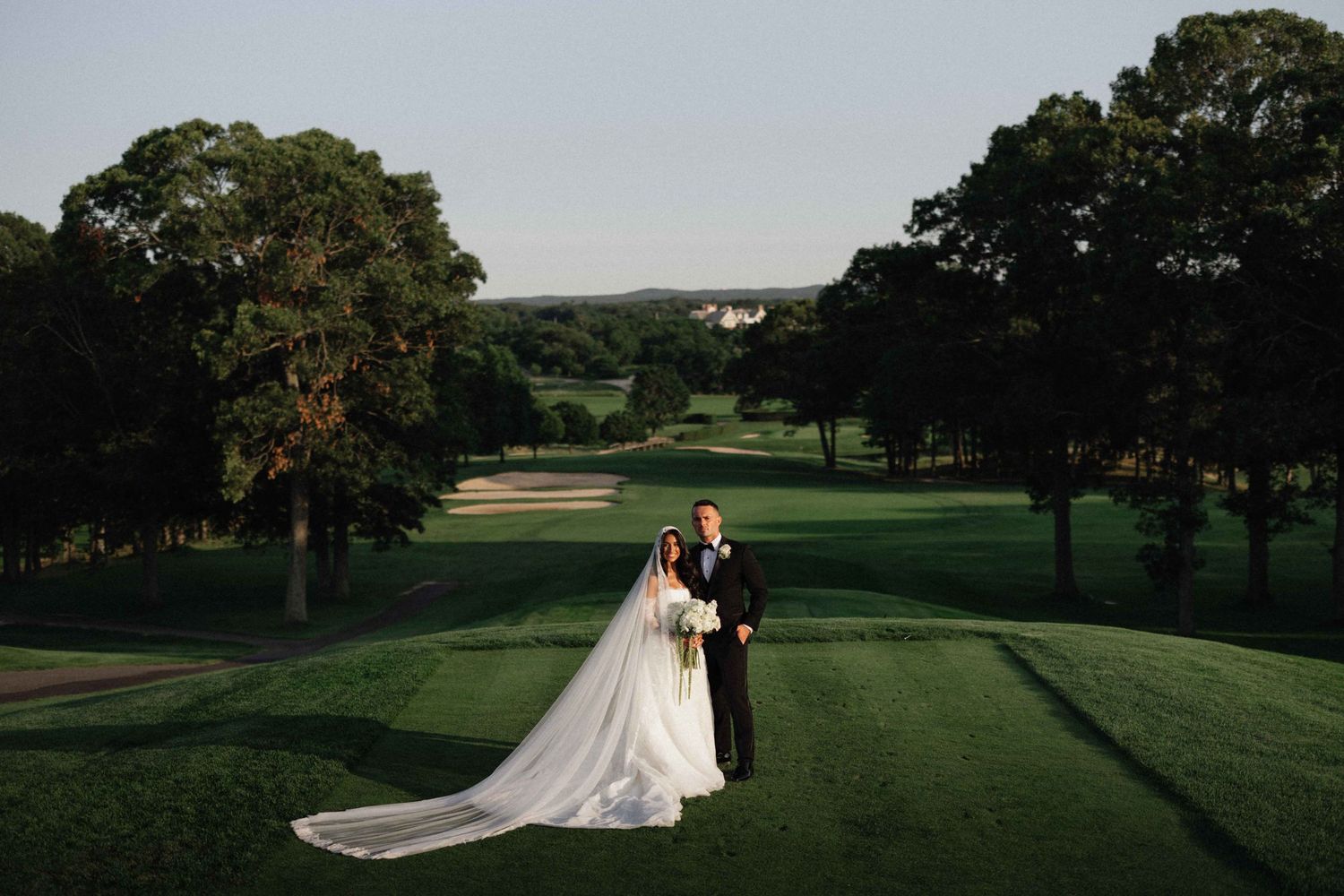 Bride and groom pose on lush green golf course with sweeping landscape and tall trees at golden hour wedding.