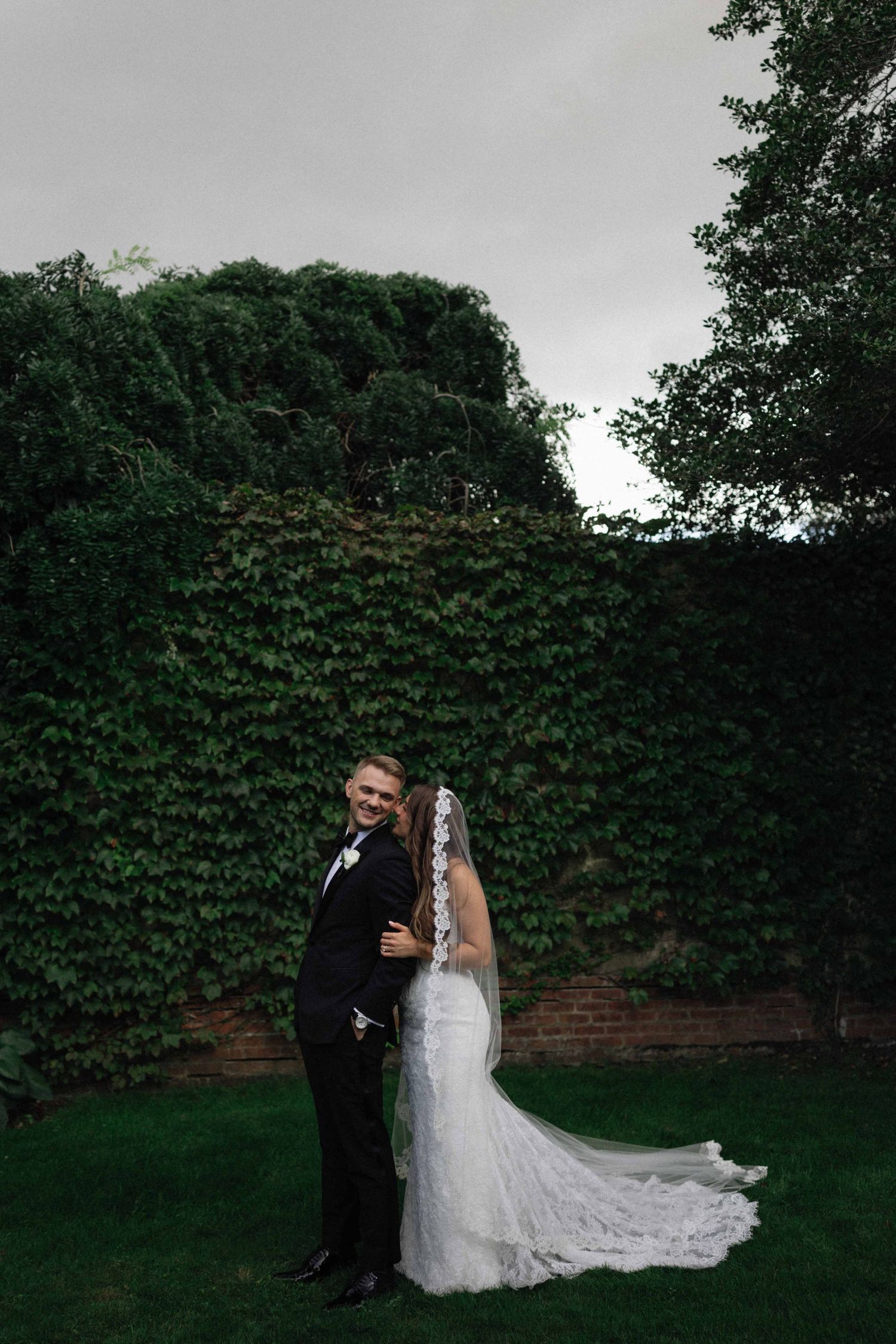 Bride and groom embracing outdoors in front of ivy-covered wall, bride in lace gown with veil.