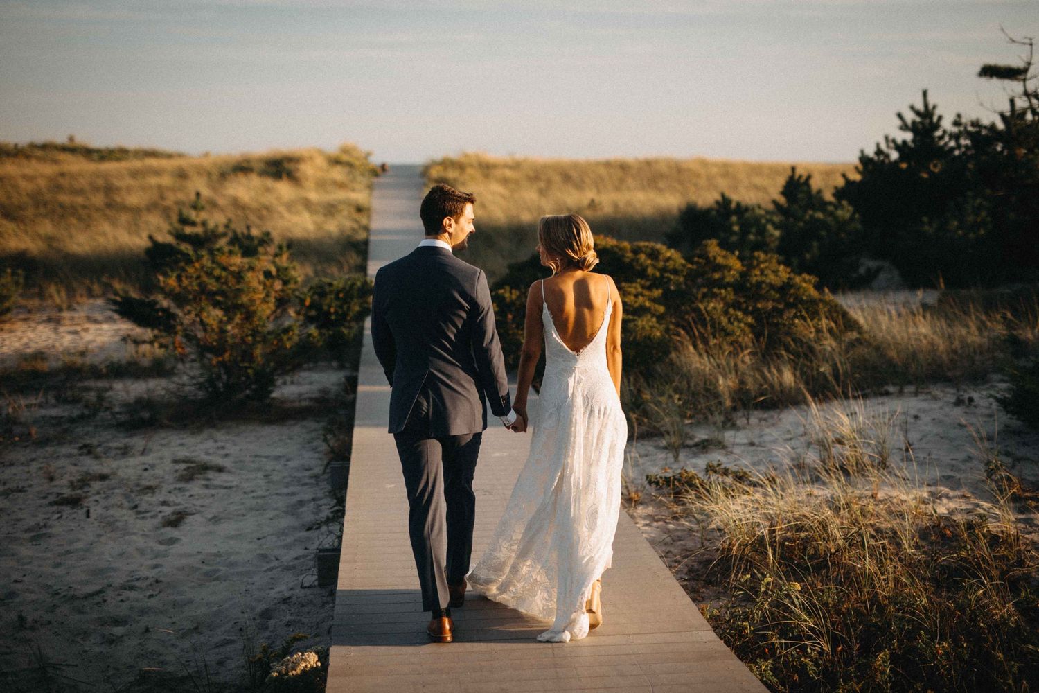 Bride and groom walk hand in hand along coastal boardwalk at golden hour sunset.