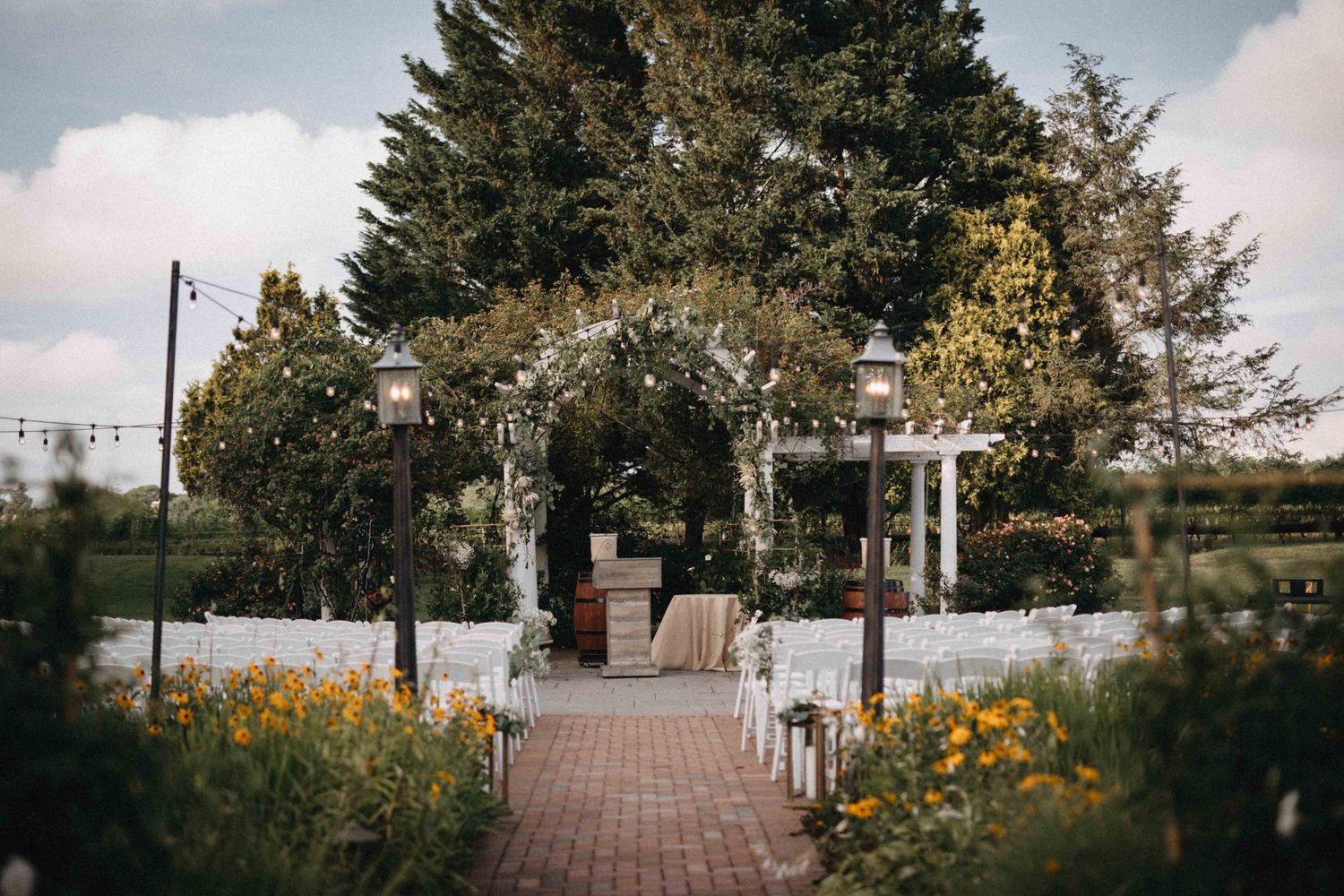 Elegant outdoor wedding ceremony aisle with floral arch, lanterns, string lights, and orange wildflowers lining brick path.