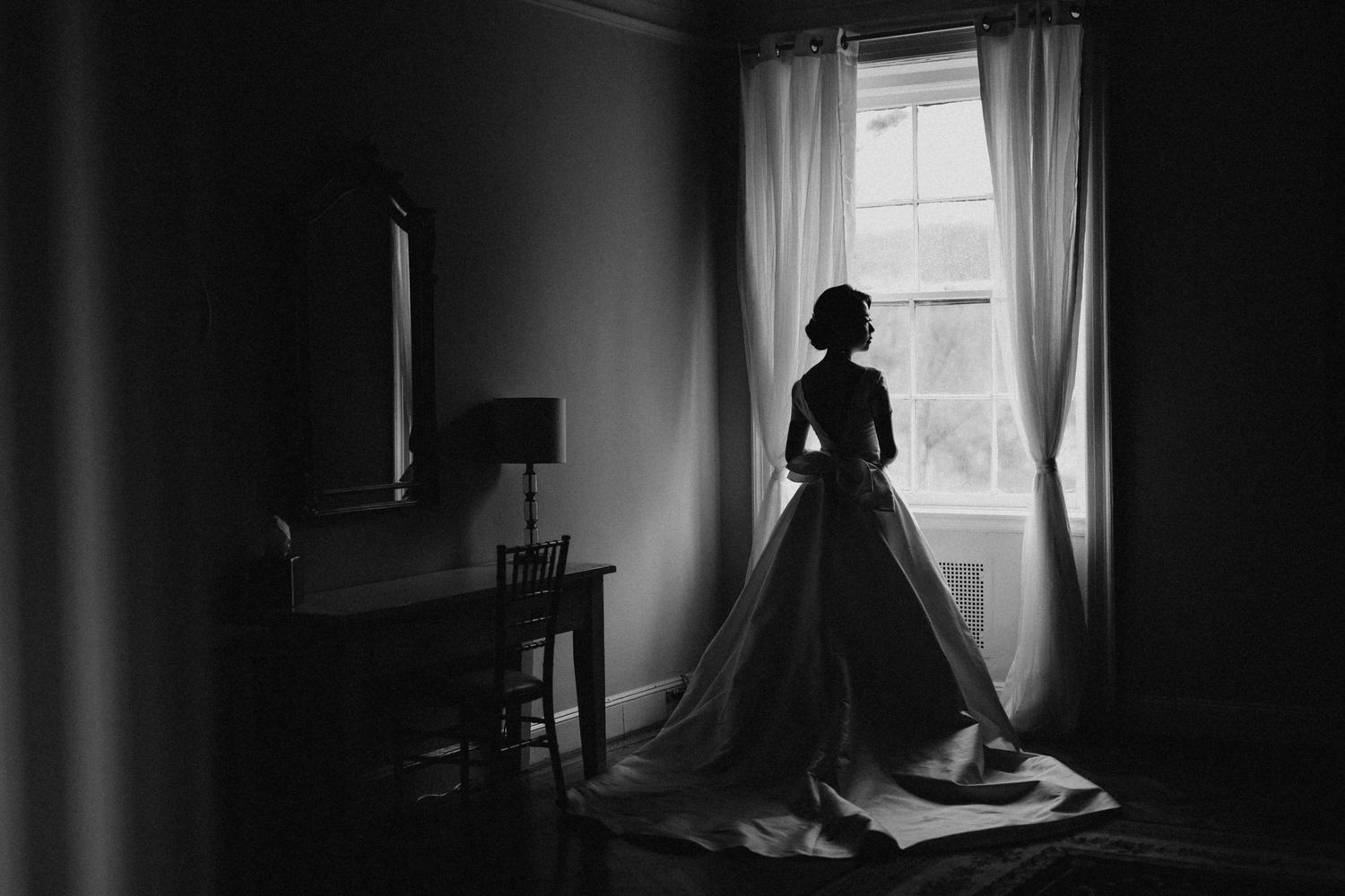 Bride in wedding gown stands silhouetted against window in black and white bridal preparation photo.