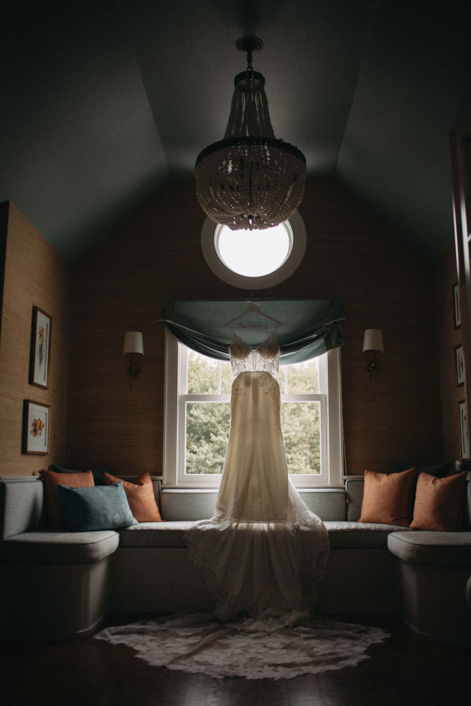 Elegant wedding dress hanging in a romantic room with chandelier and circular window.