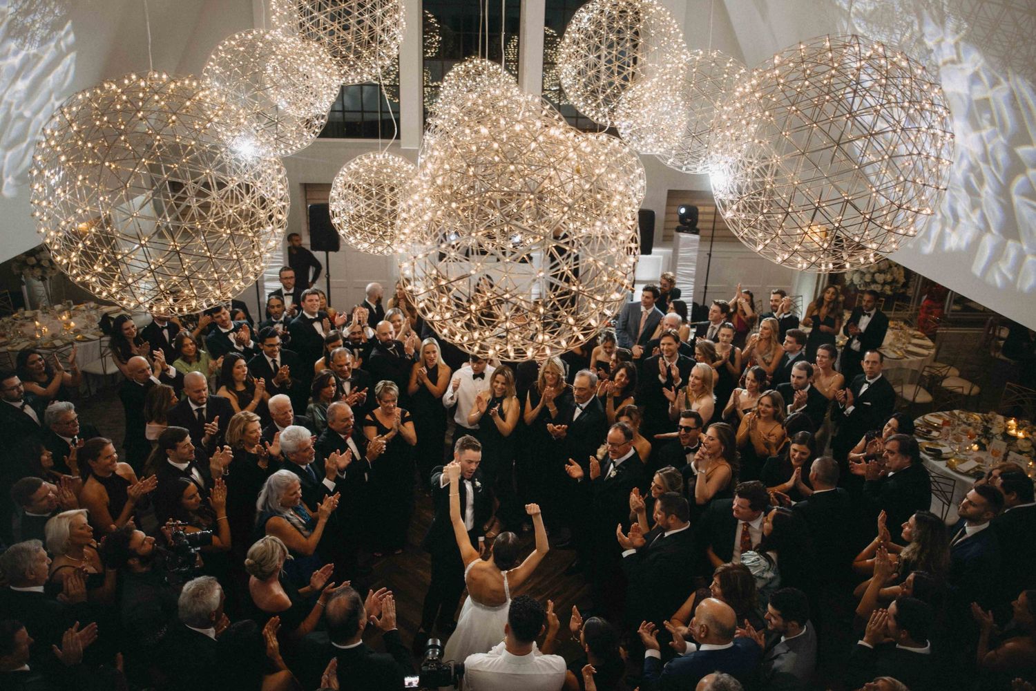 Aerial view of bride and groom celebrating with guests under dazzling spherical chandeliers.