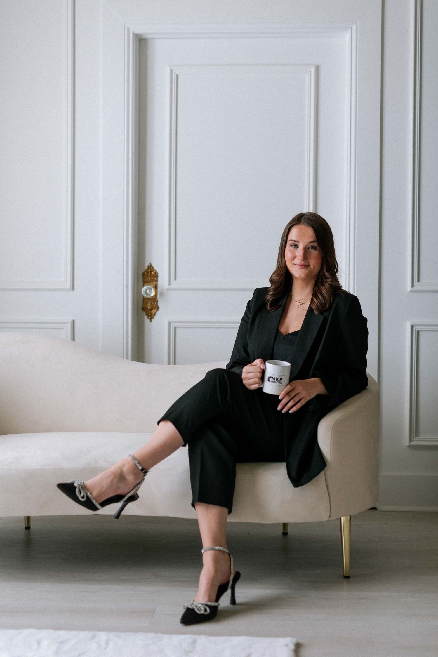Confident brunette woman in black suit sitting on cream sofa holding white coffee mug.