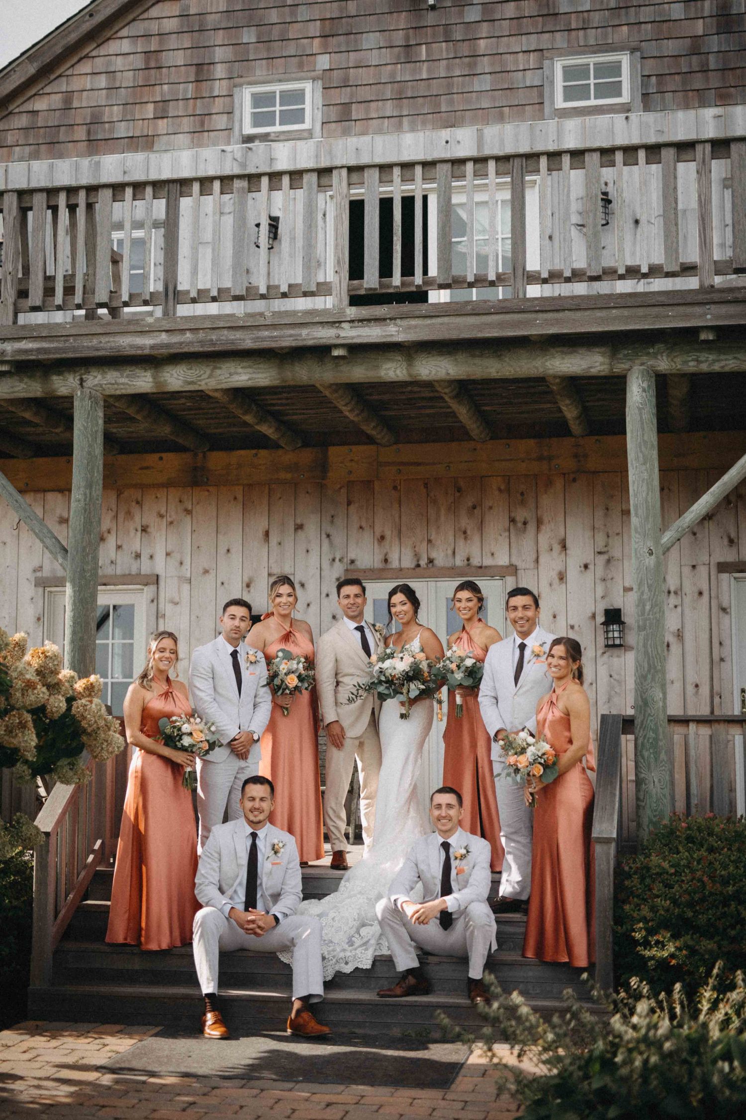 Wedding party poses on barn steps, bridesmaids in coral gowns and groomsmen in white shirts.