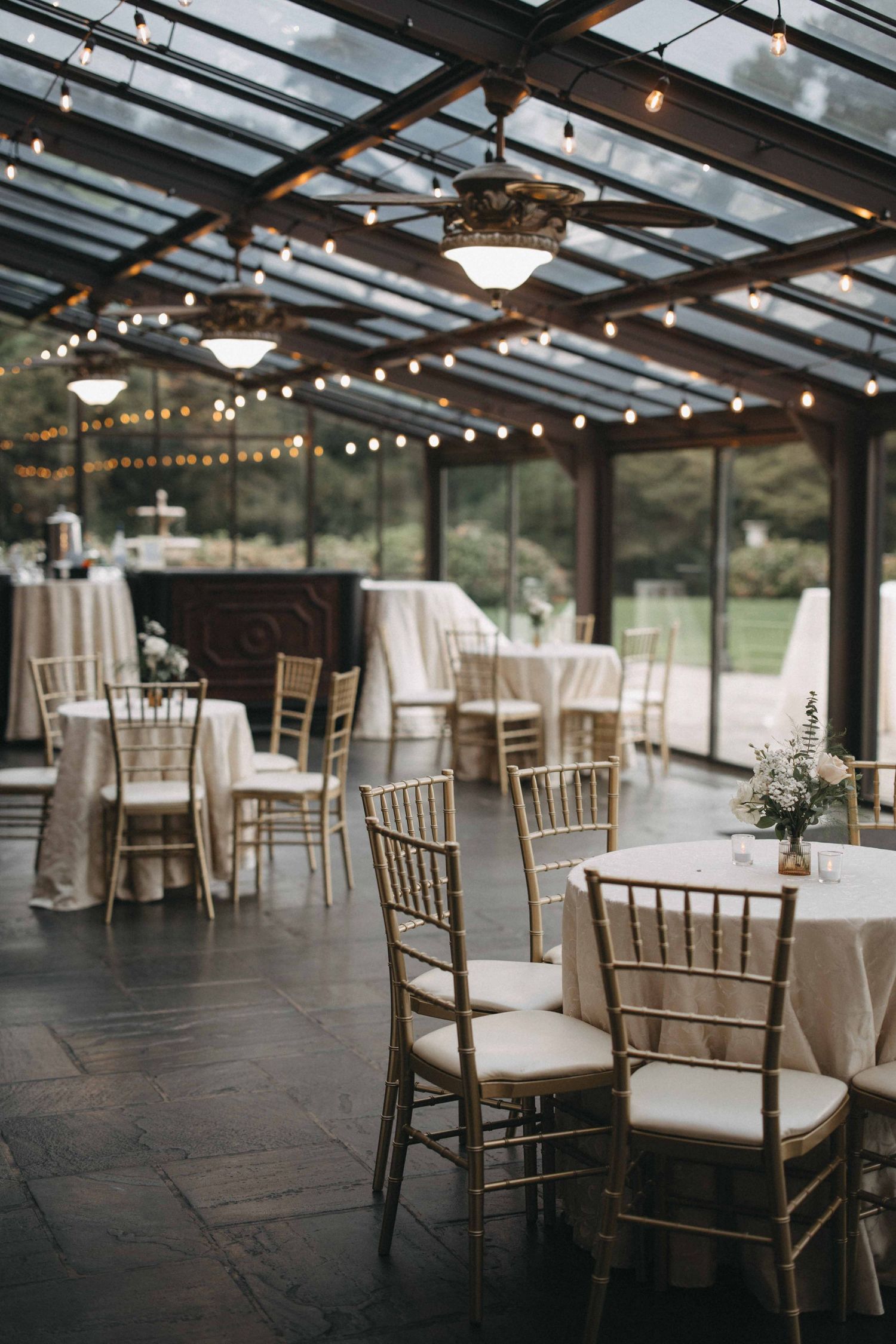 Elegant wedding reception in a glass conservatory with string lights, gold chiavari chairs, and white linen-draped round tables.