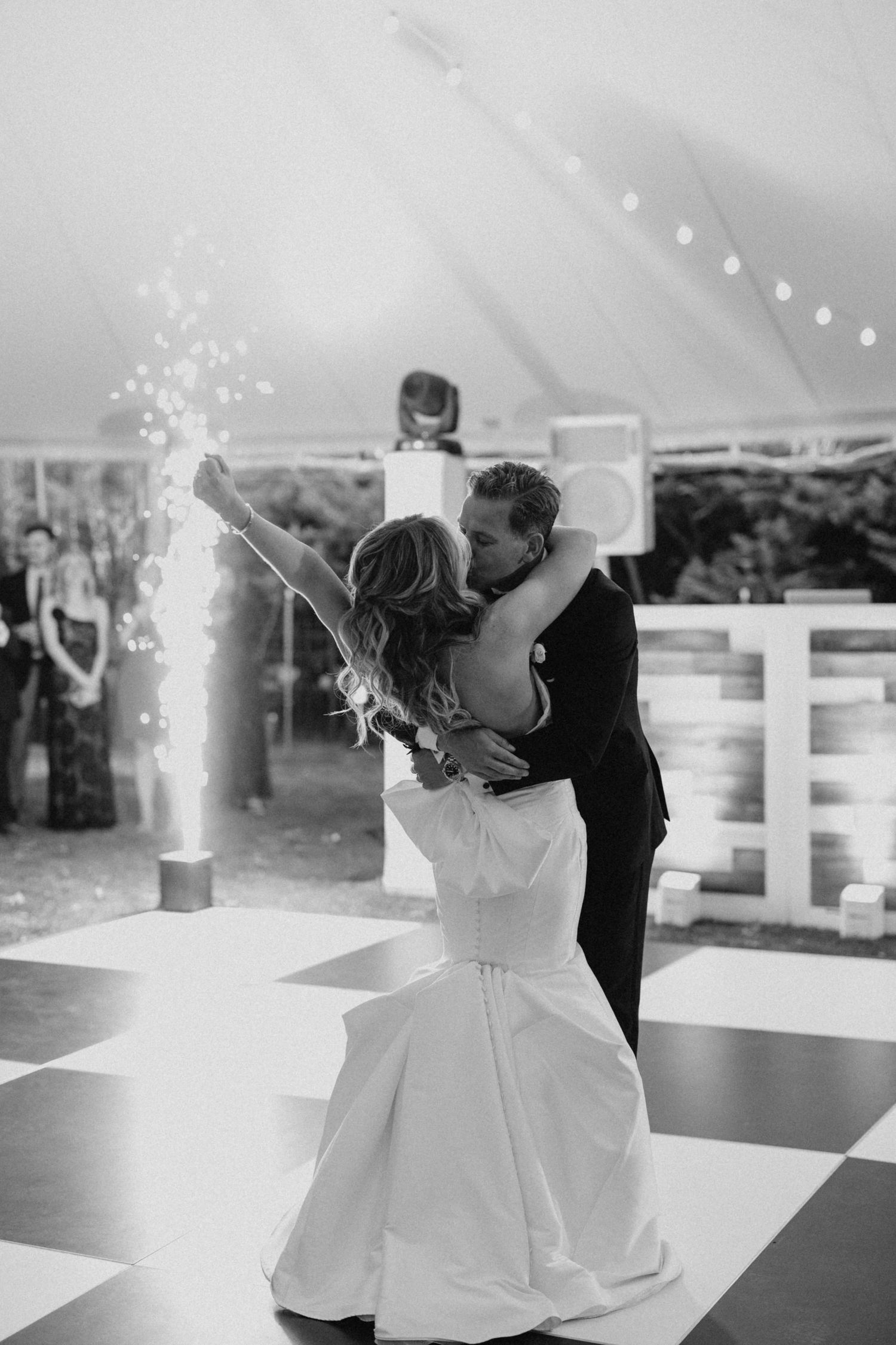 Black and white photo of bride and groom sharing first dance with sparklers on checkered dance floor.
