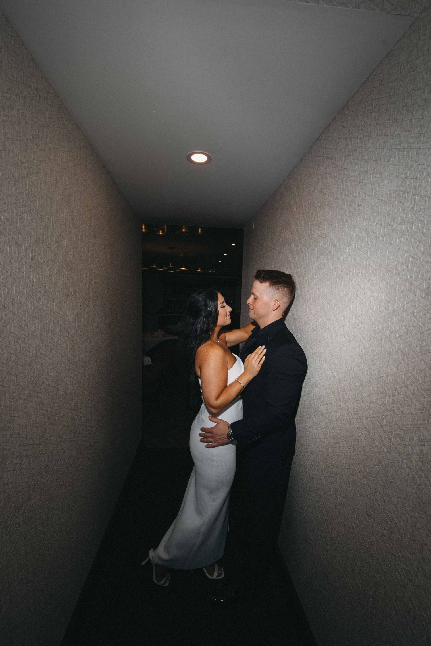 Couple in elegant attire sharing intimate moment in narrow dimly lit hallway, woman in white dress.