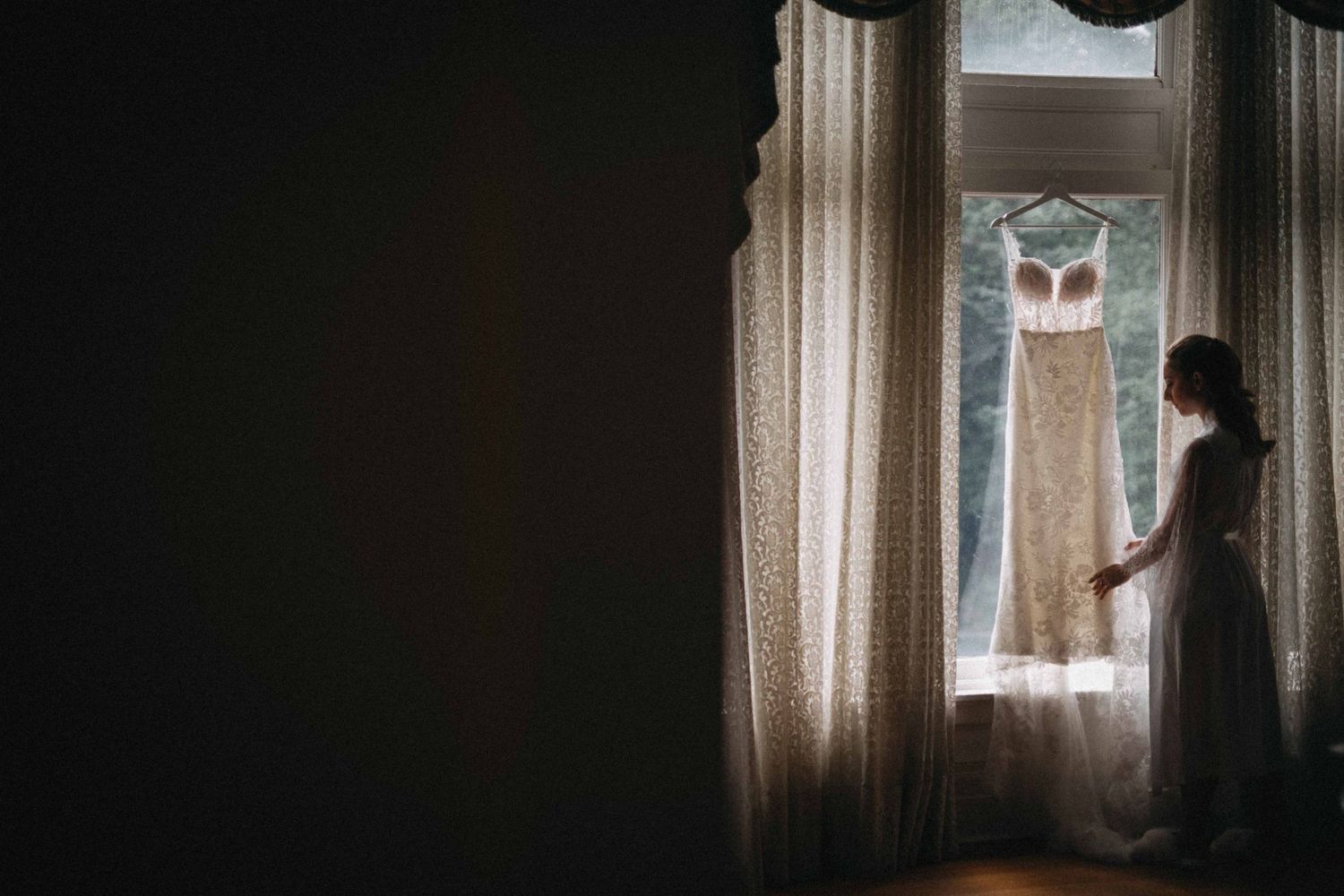 Bride silhouetted beside window with elegant lace wedding dress hanging in soft natural light.