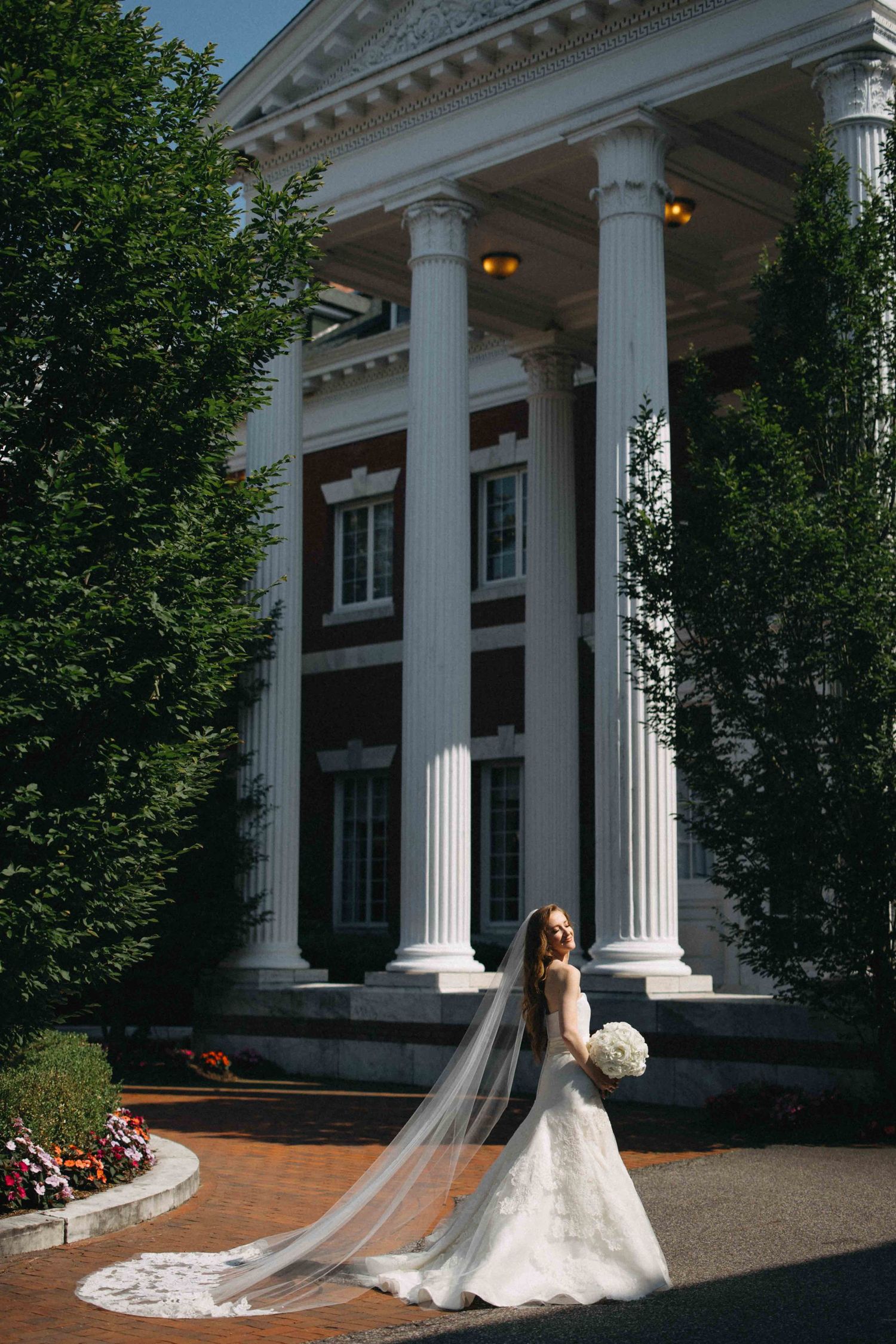 Bride in lace gown with cathedral veil poses before grand colonial mansion with white columns.