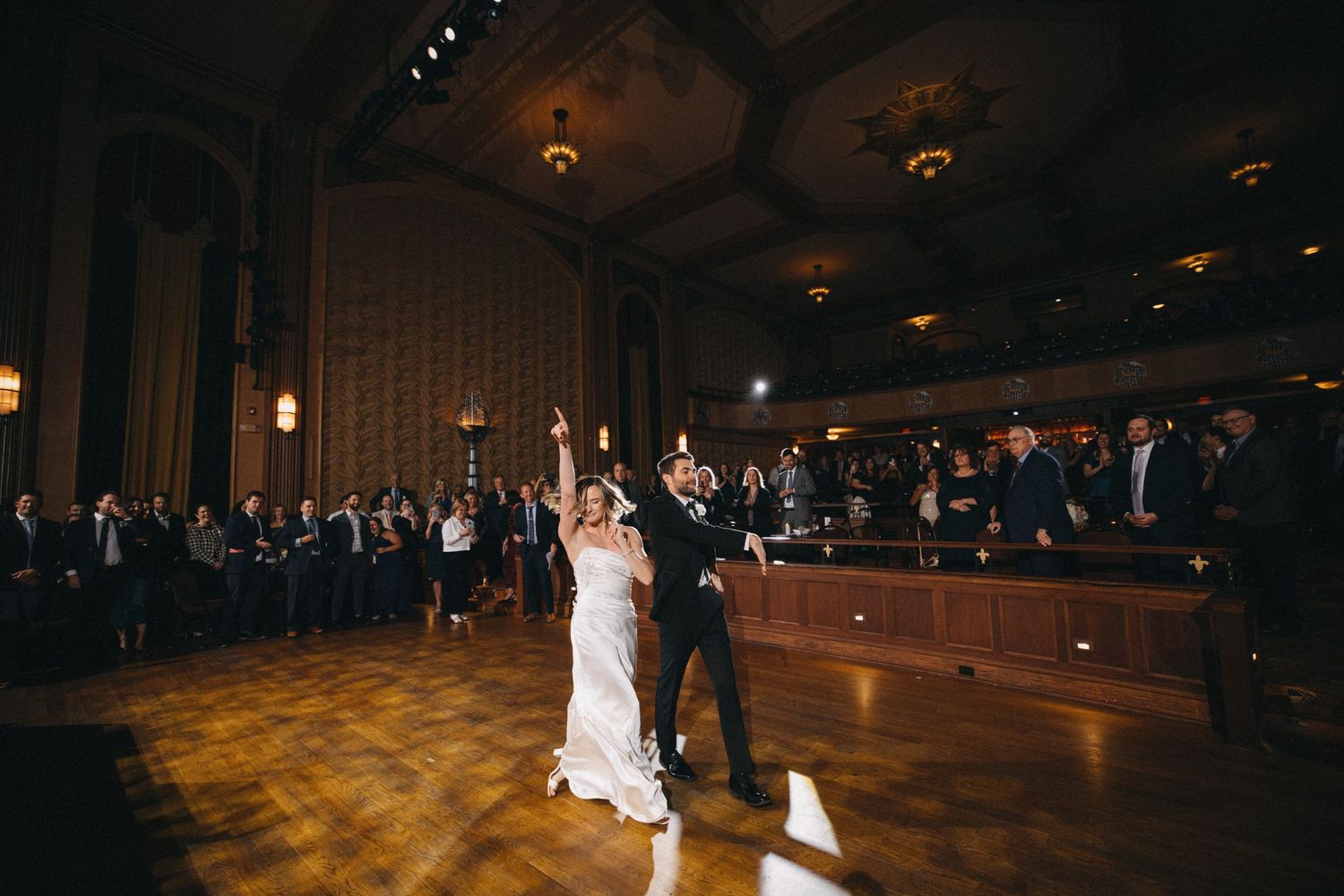 Bride and groom celebrate first dance on wooden ballroom floor surrounded by wedding guests in grand venue.