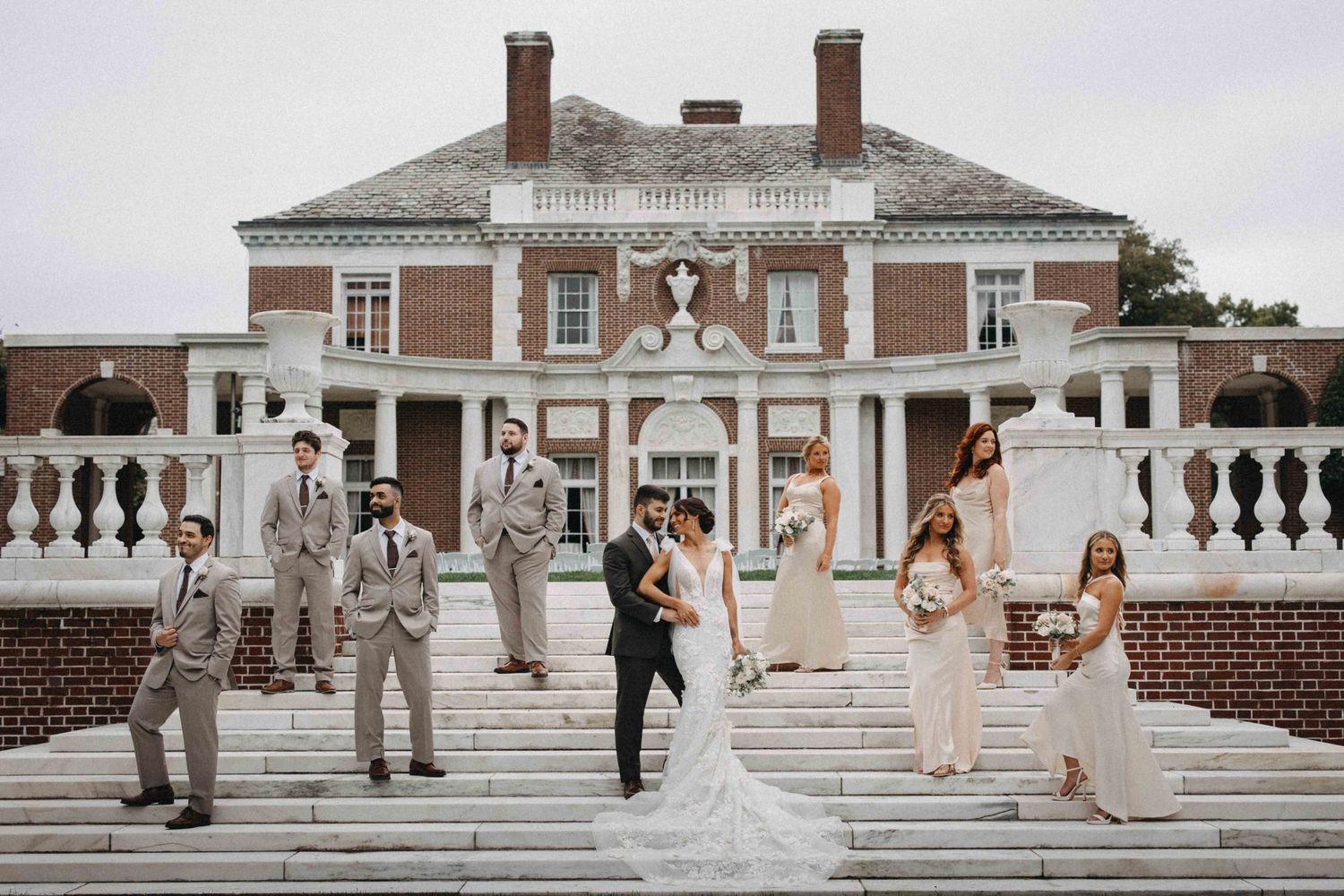 Wedding party poses on grand stone steps of elegant brick Georgian manor house on overcast day.