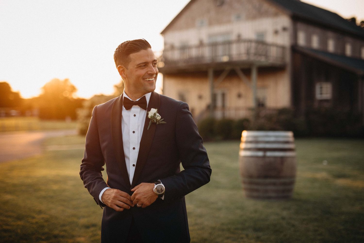 Dapper groom in navy tuxedo smiles at sunset outside rustic barn wedding venue.