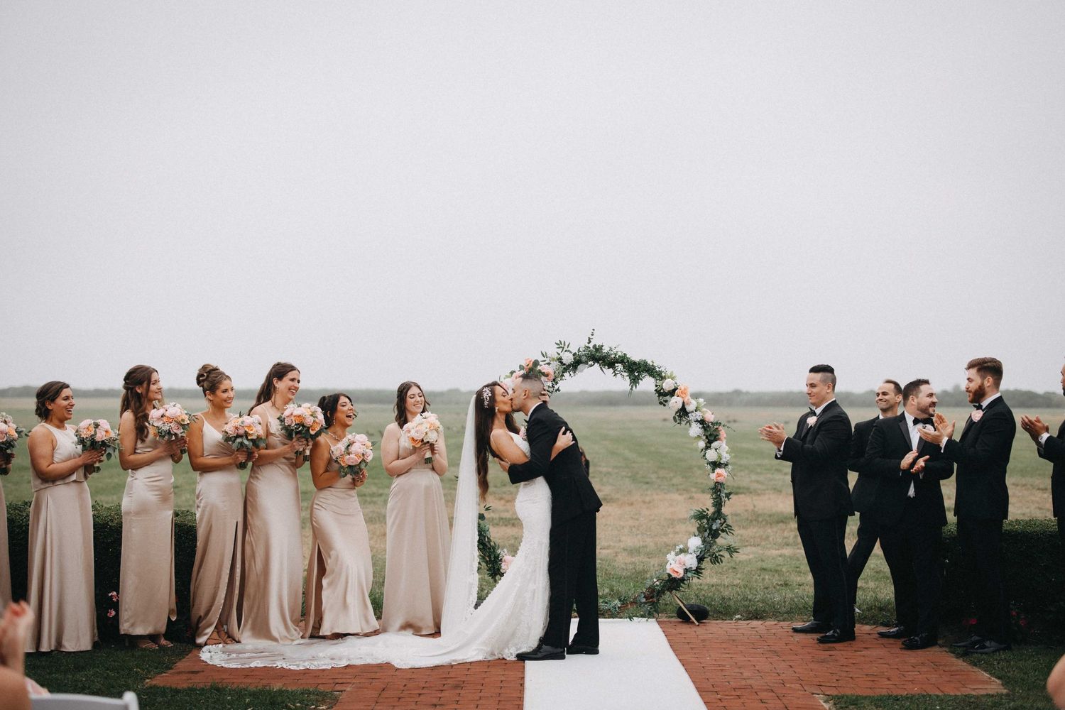 Bride and groom share first kiss at altar with bridesmaids and groomsmen at outdoor ceremony.