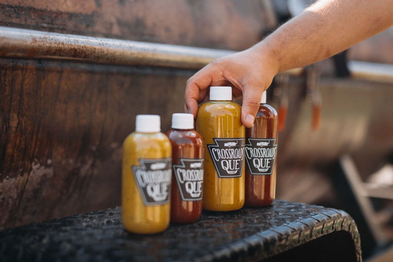 Hand reaching for Cotton Gin Que BBQ sauce bottles displayed on rustic smoker surface.