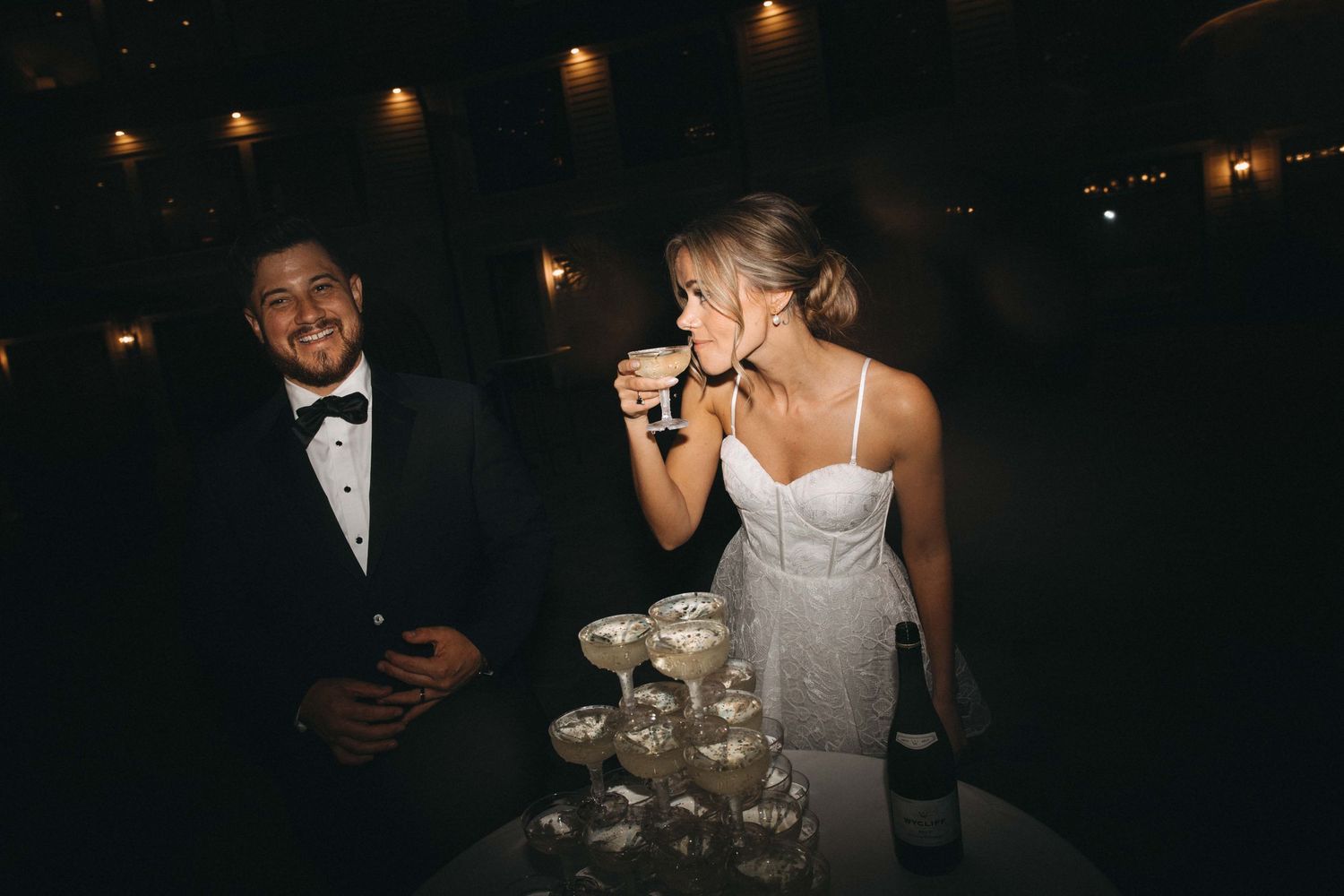 Bride sipping champagne beside groom at elegant wedding champagne tower display.