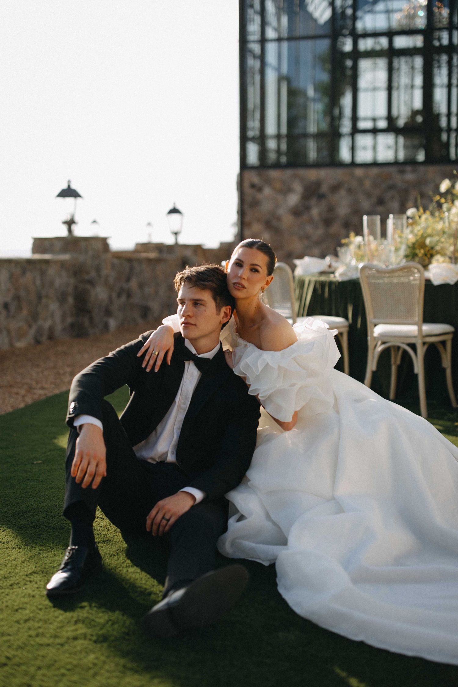 Bride in white gown and groom in black tuxedo sitting together on green grass at elegant outdoor wedding venue.