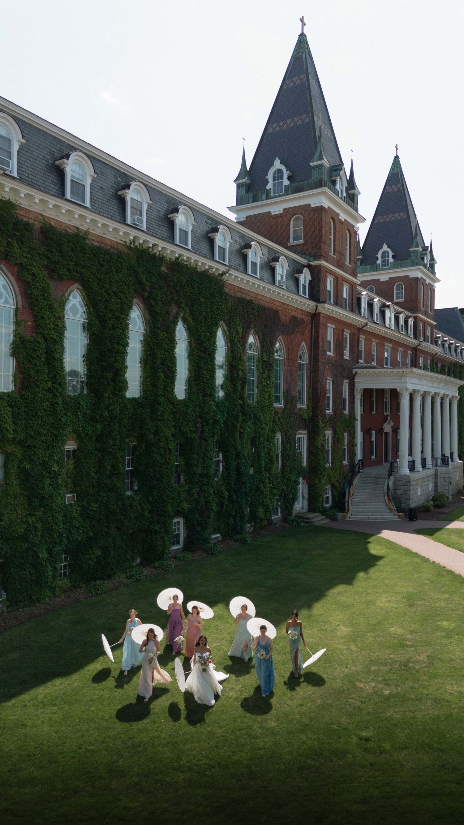 Wedding party with parasols on lawn of ivy-covered red brick Gothic university building with dark spires.