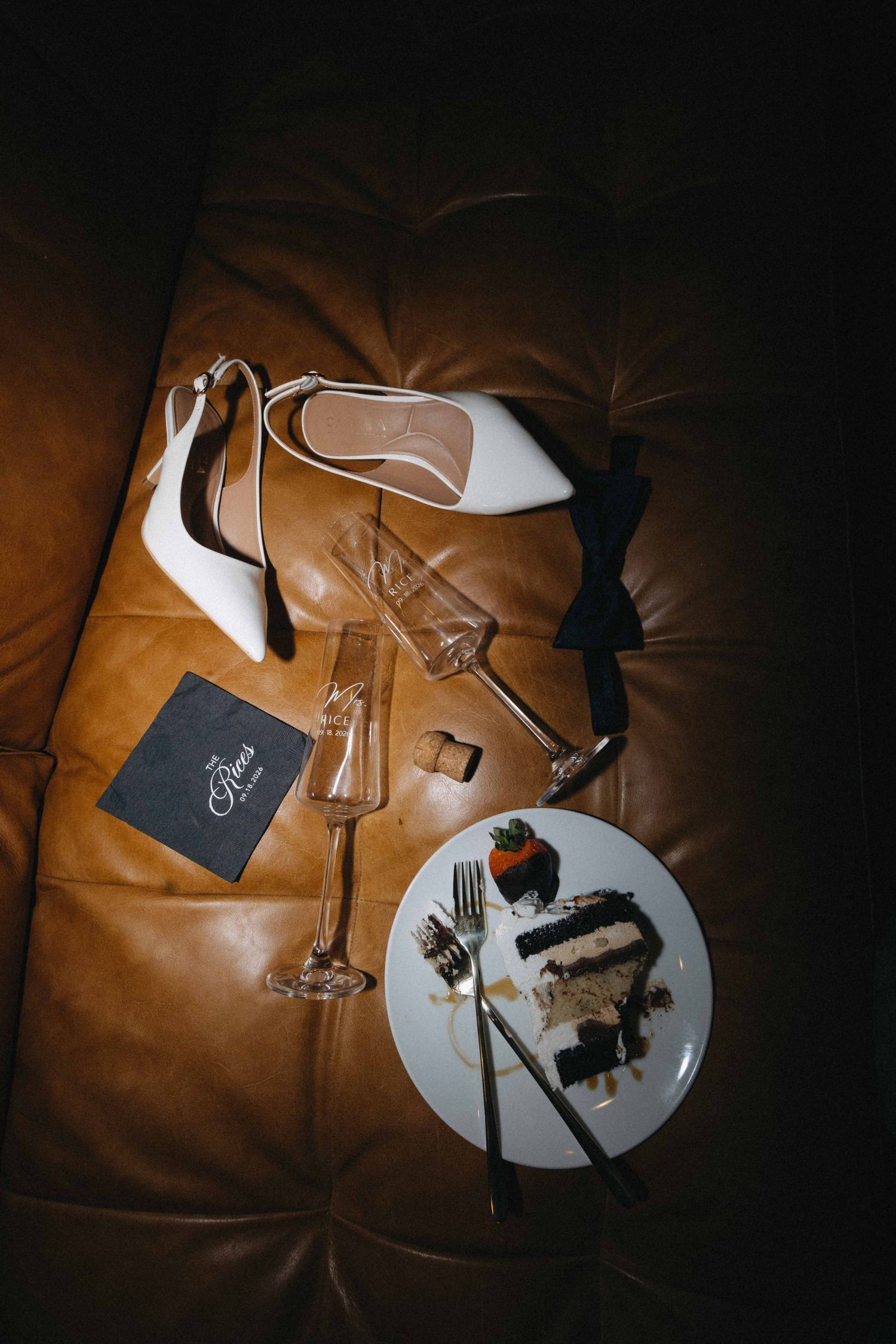 Elegant wedding flat lay with white heels, bow tie, champagne glasses, cake slice, and menu on brown leather sofa.