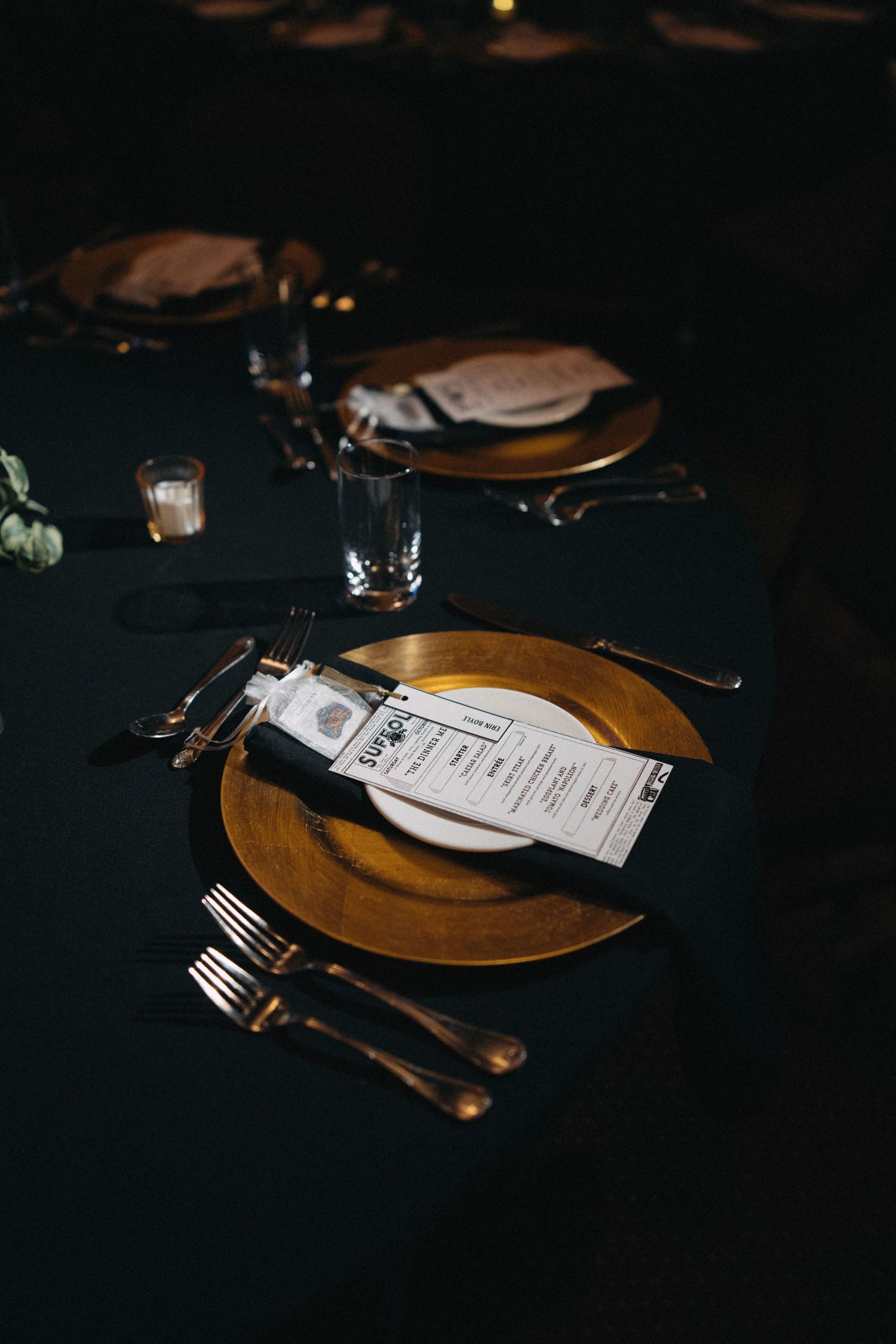 Elegant wedding table setting with gold charger plates, dark linens, and printed menu cards.