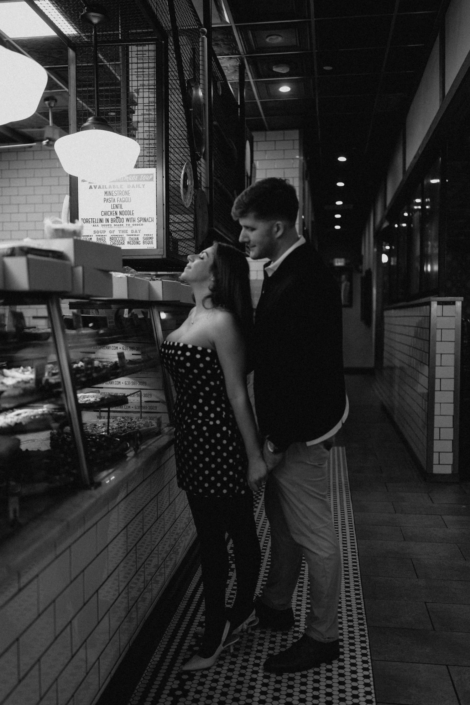Black and white photo of couple sharing romantic moment inside a deli or bakery shop at night.