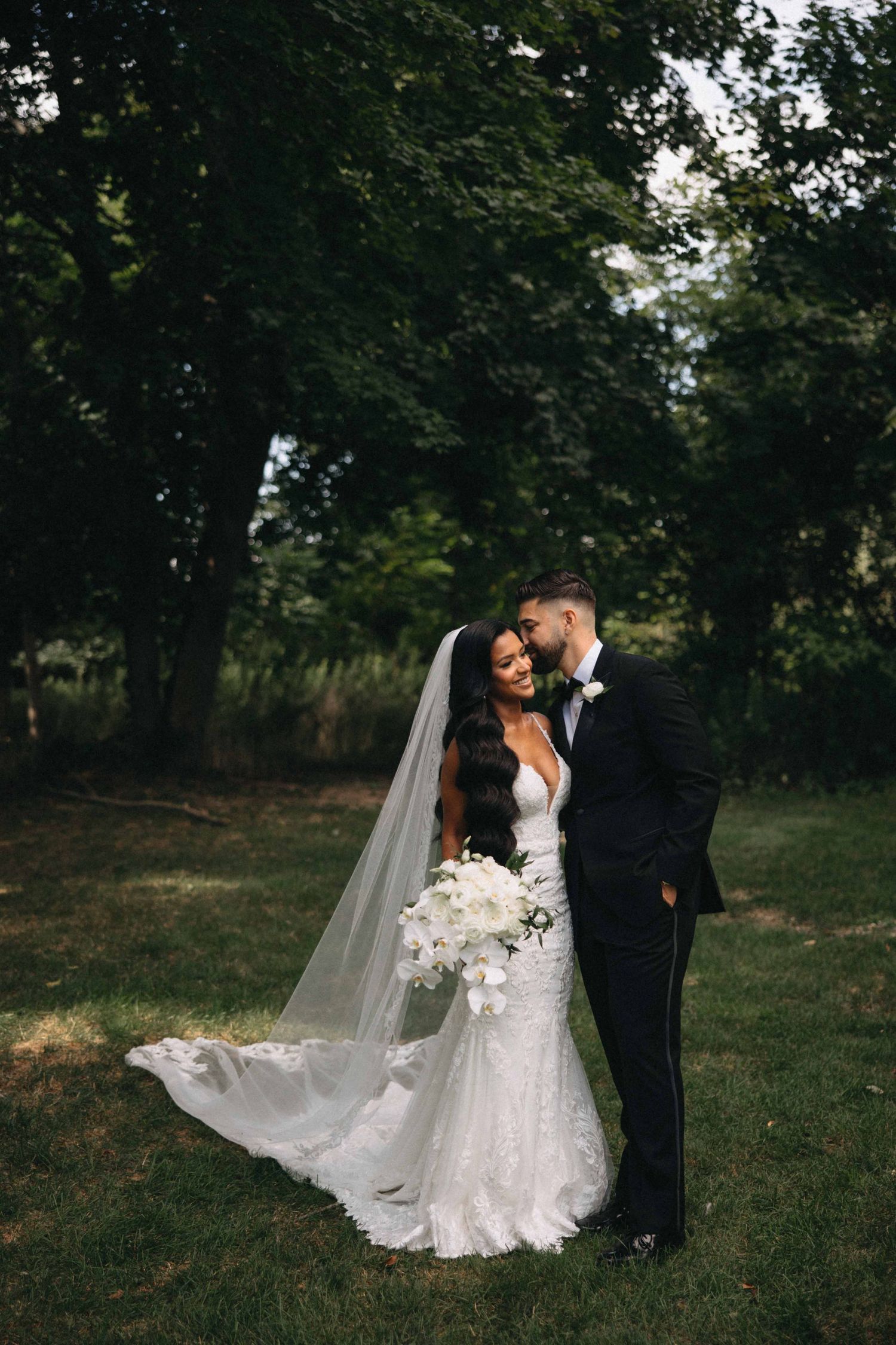 Bride in lace mermaid gown with cathedral veil and bouquet embracing groom outdoors.
