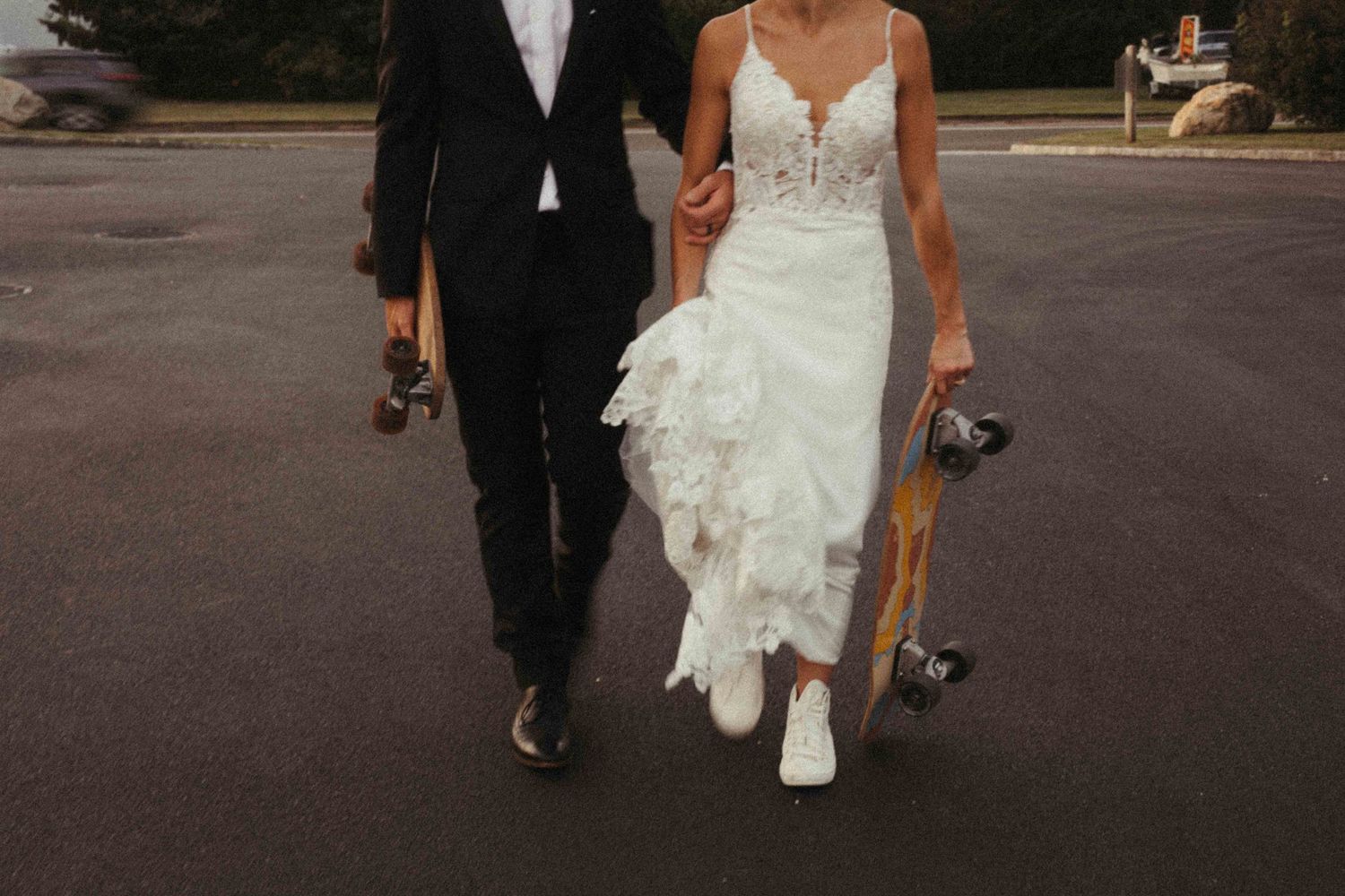 Bride and groom walking in a parking lot carrying longboards in wedding attire.