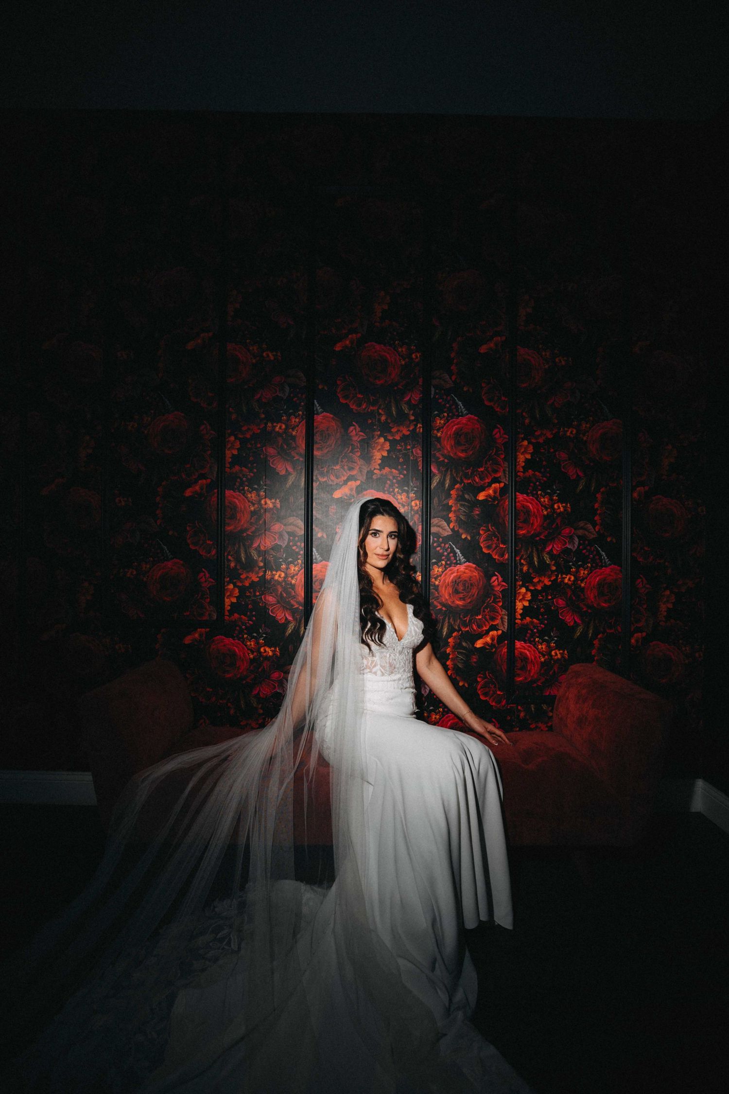 Elegant bride in white gown and long veil seated before dramatic dark floral wallpaper backdrop.