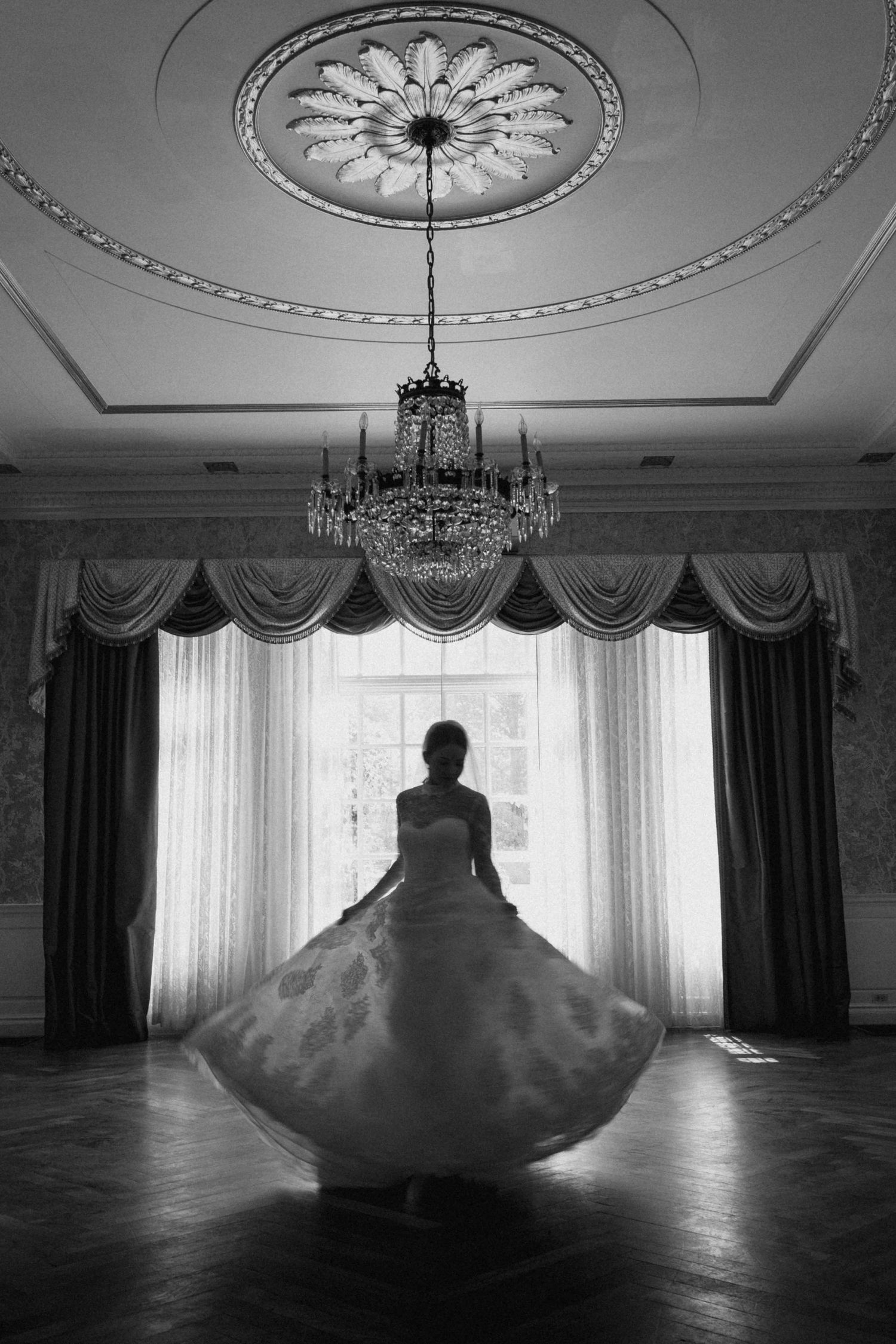 Black and white photo of a bride twirling in a ballroom with chandelier and ornate ceiling.