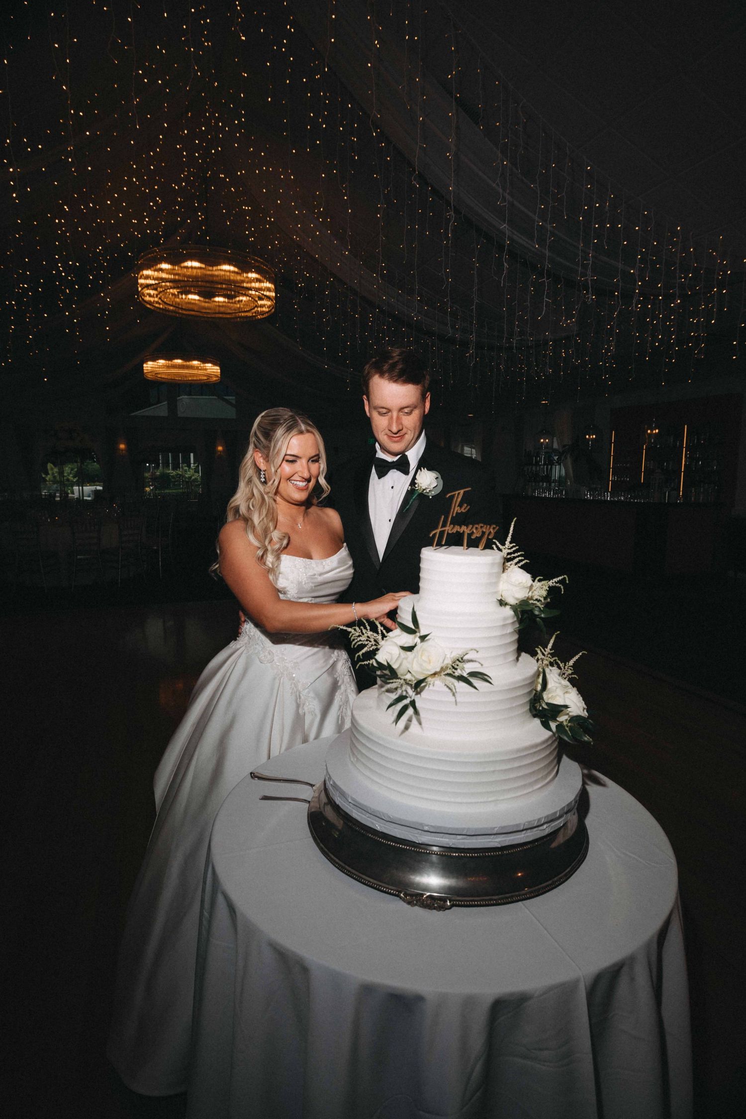 Bride and groom cutting a white three-tier wedding cake decorated with greenery under a starlit ceiling.