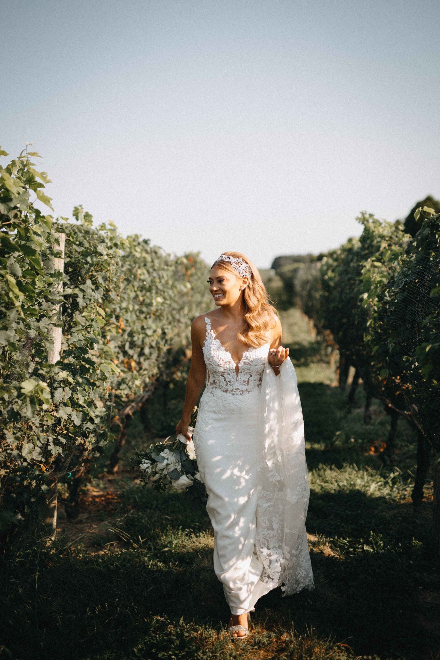 Smiling bride in lace V-neck wedding gown walking through lush vineyard rows on wedding day.