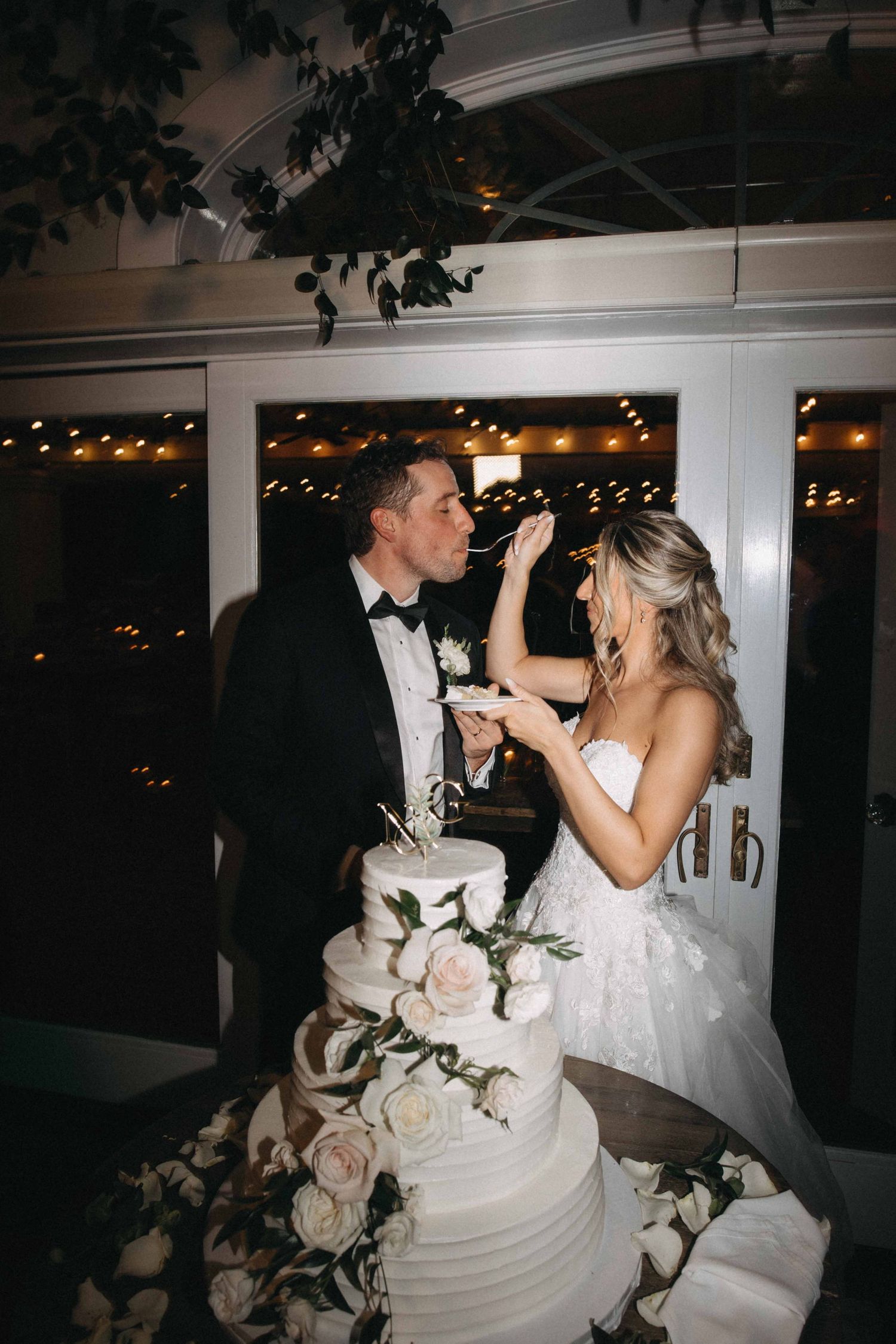 Bride feeding groom a slice of four-tier white wedding cake decorated with roses at evening reception.
