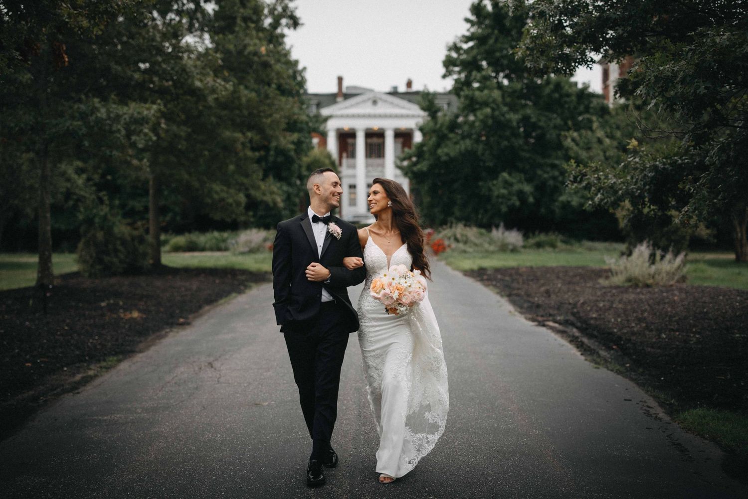 Bride and groom walk hand in hand down a tree-lined path toward a white colonial mansion.
