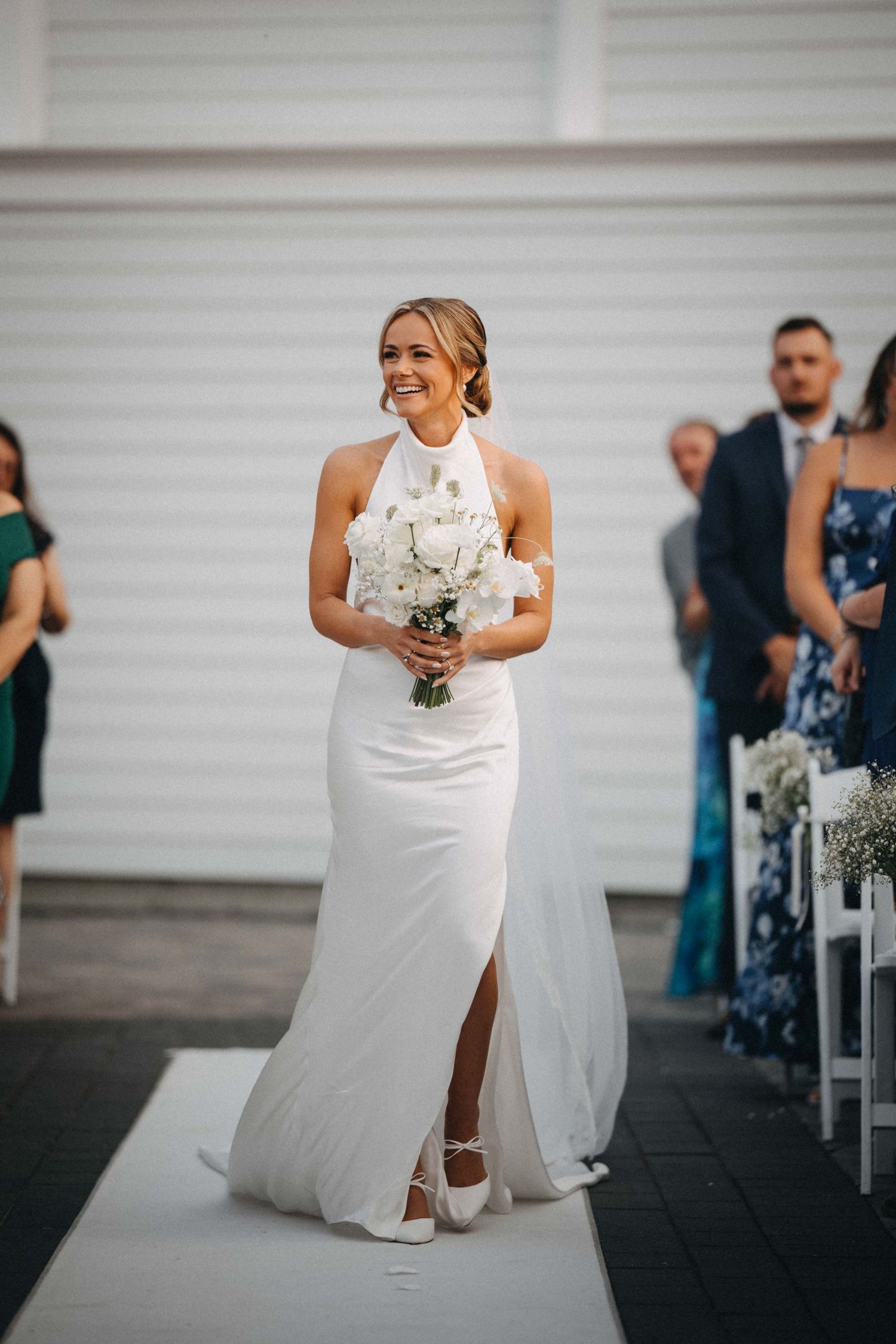 Smiling bride walks down the aisle in a white halter gown holding a white floral bouquet.