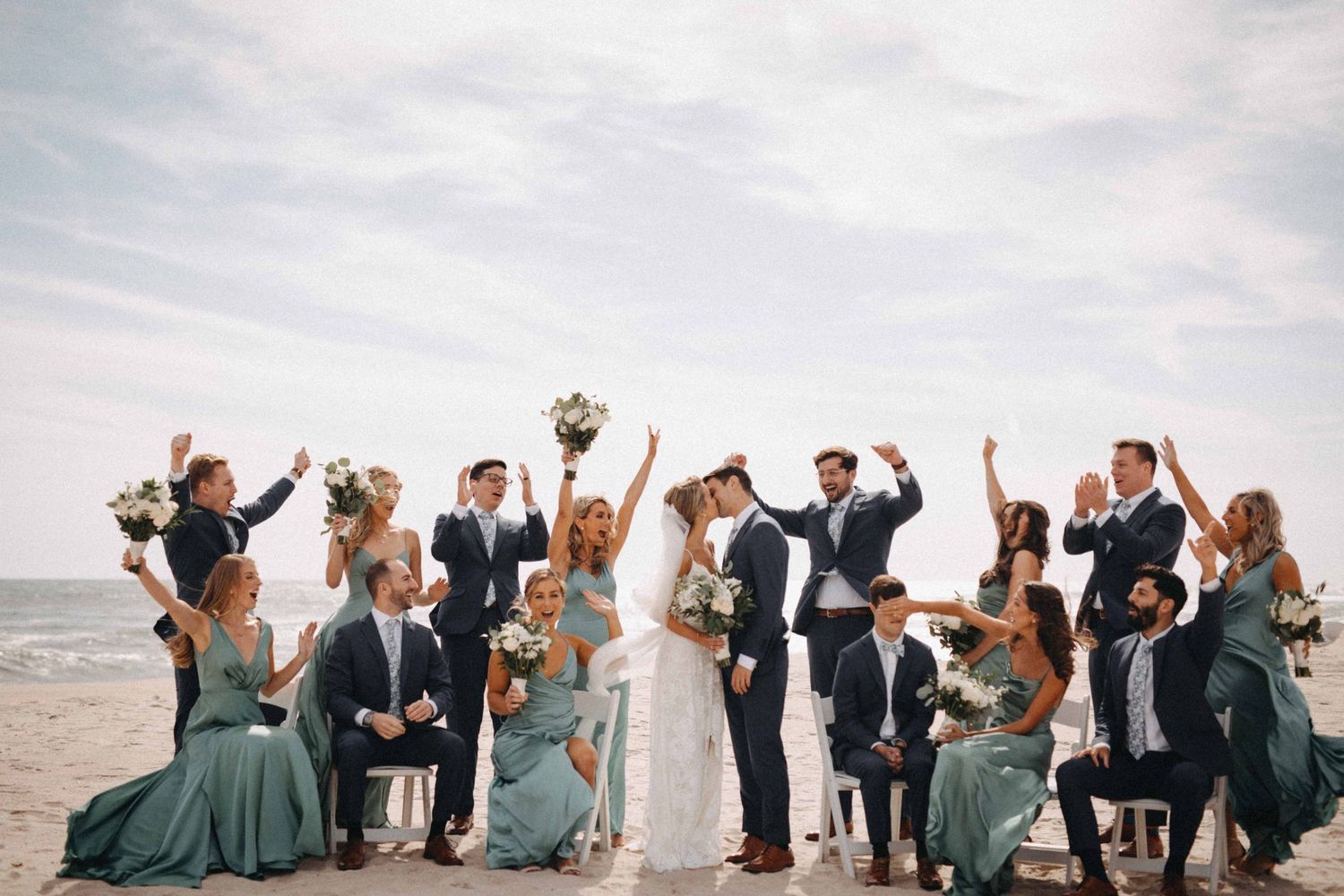 Wedding party celebrates on beach with bridesmaids in teal gowns and groomsmen in navy suits.